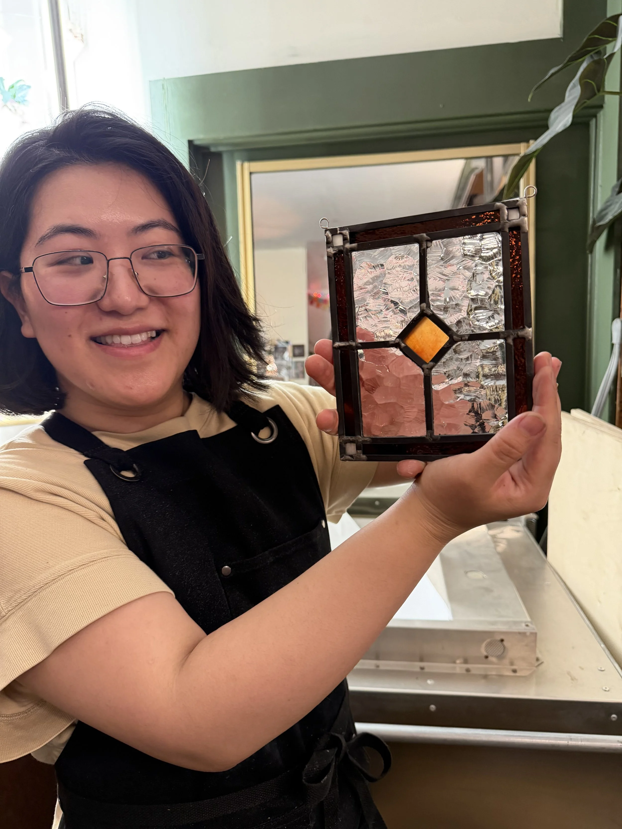 A woman with glasses smiling while holding a stained glass lantern in a room with green walls and a window in the background.