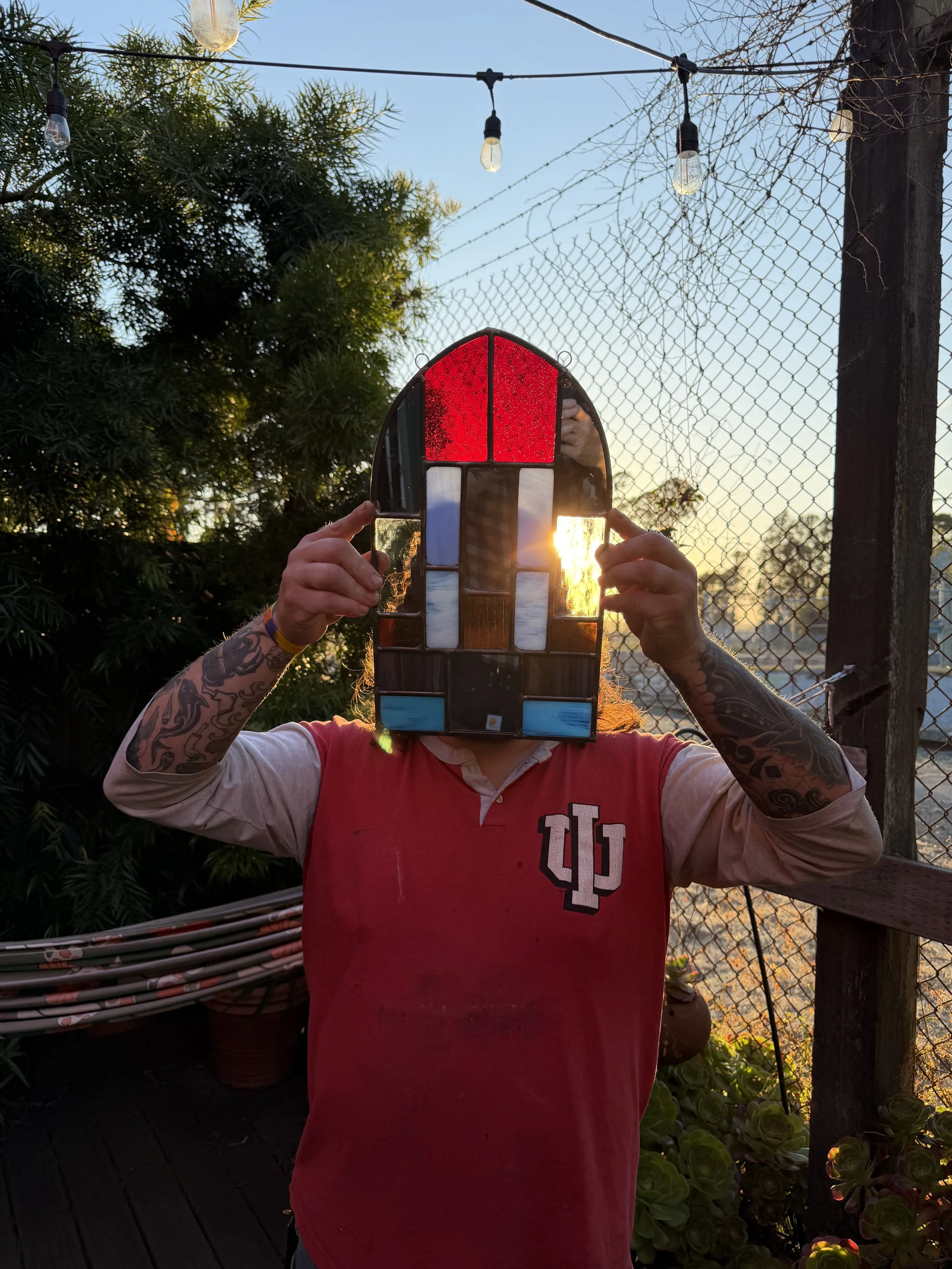 Person holding a stained glass window with the sun shining through it during sunset, outdoors, with trees, a chain-link fence, string lights, and plants in the background.