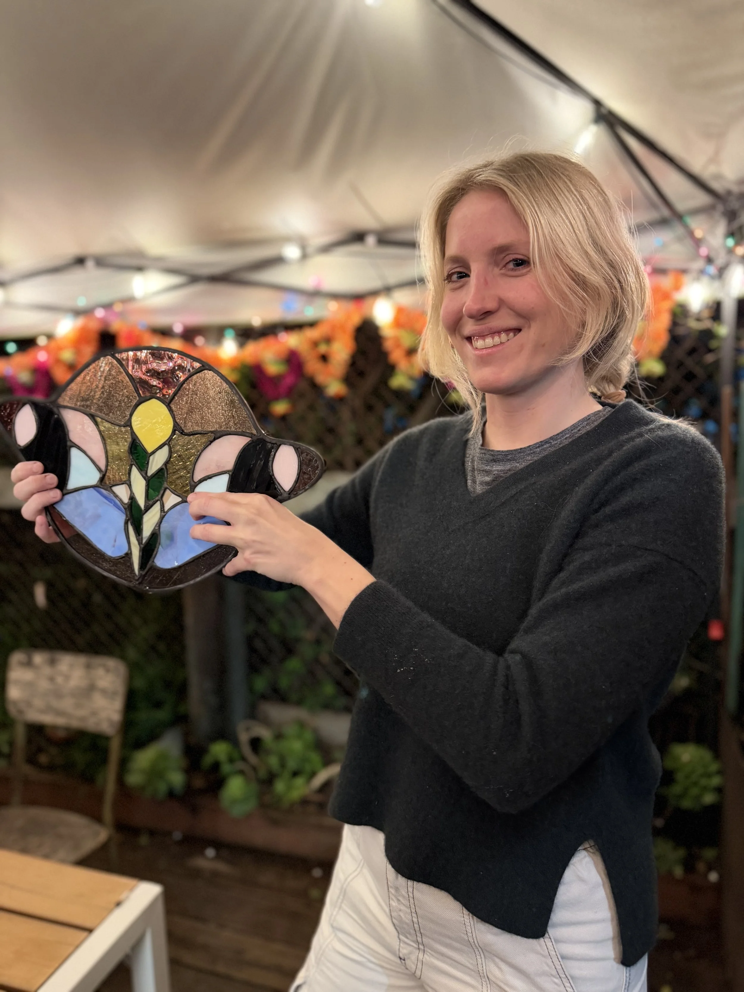 A smiling woman with blonde hair holding stained glass art in an outdoor setting decorated with string lights and colorful flowers.