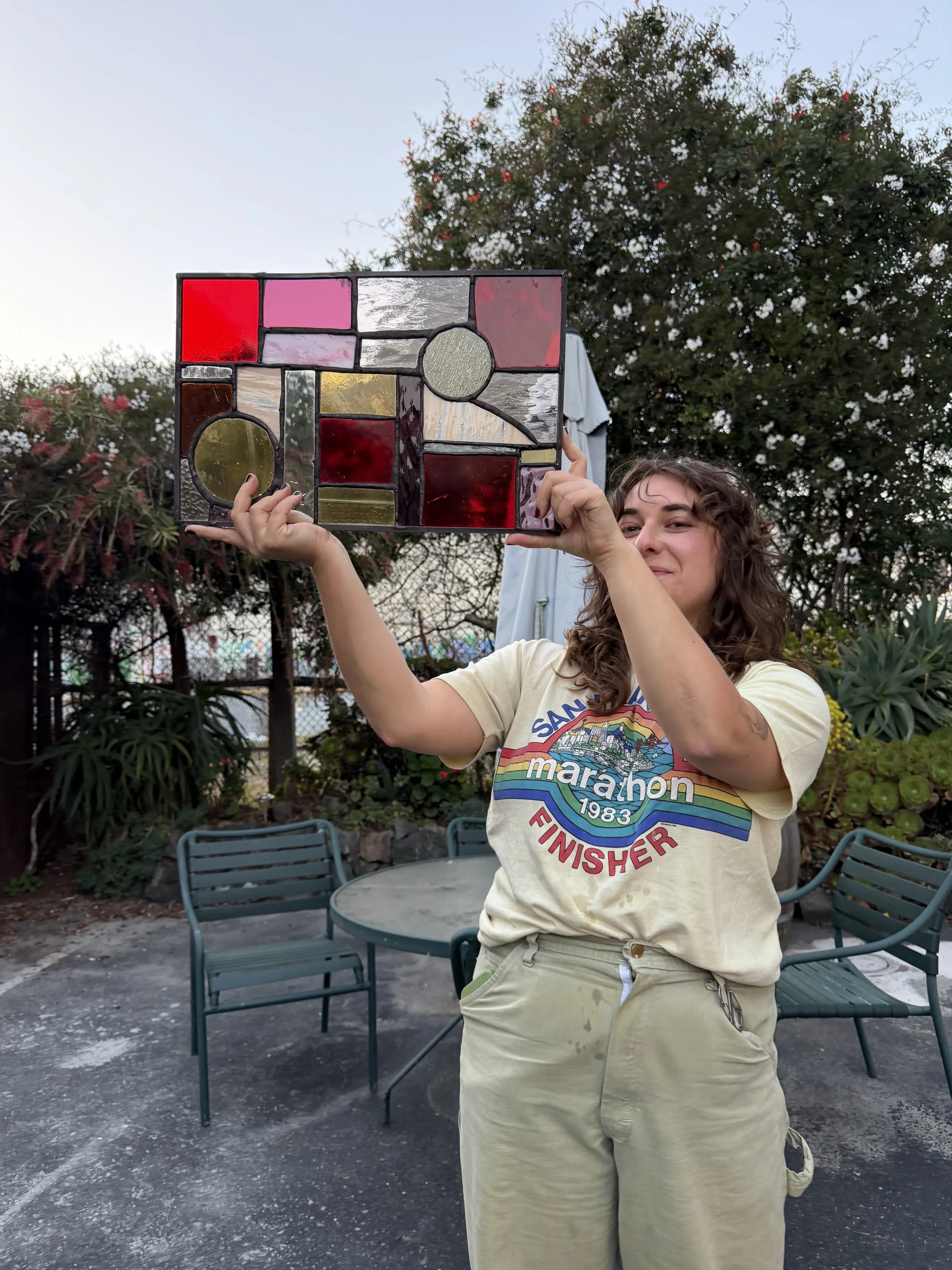 A person holding a stained glass artwork outdoors on a patio with chairs, trees, and a fence in the background.