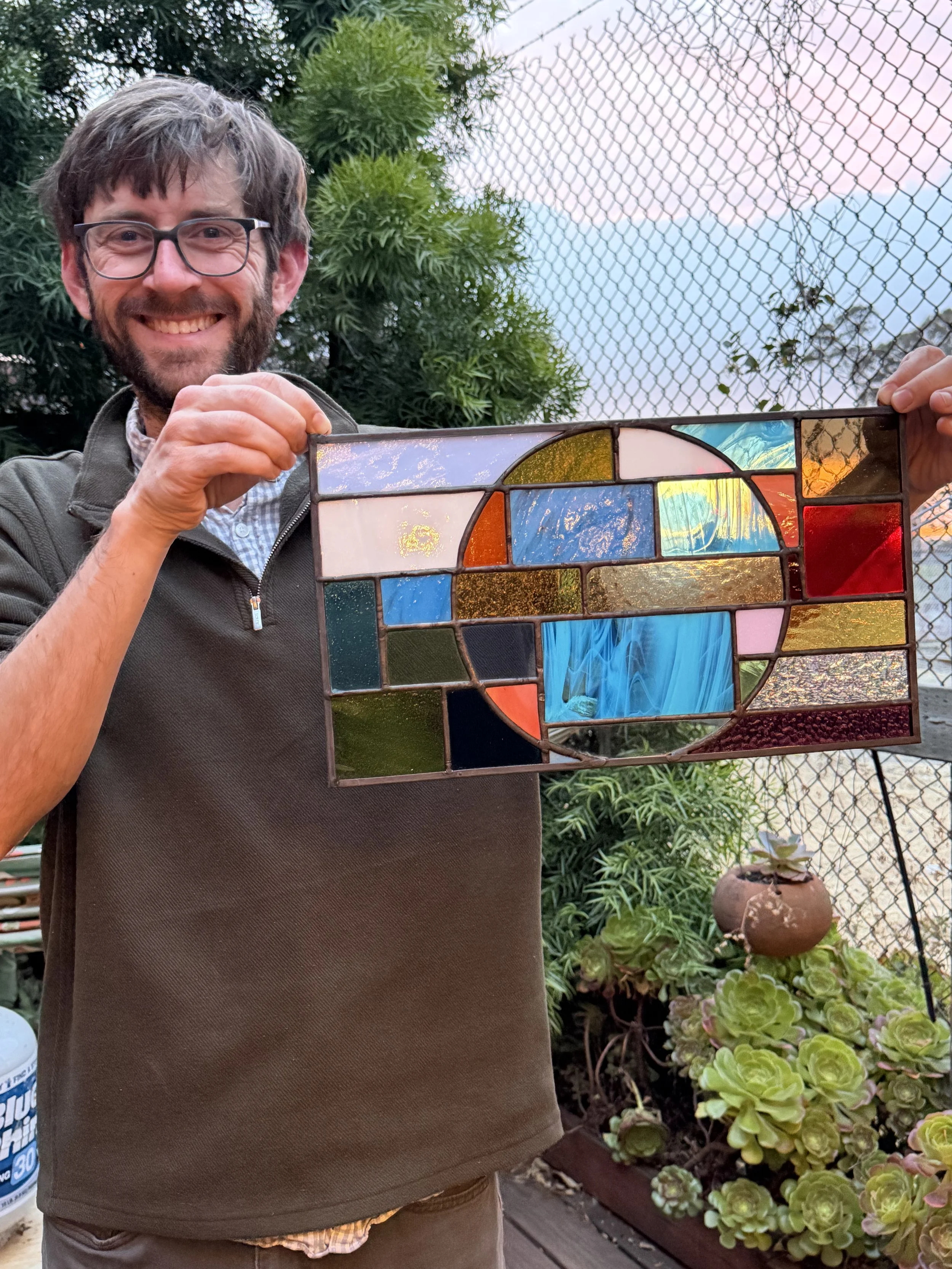 A man with glasses and a beard smiling while holding a colorful stained glass art piece in an outdoor garden setting.