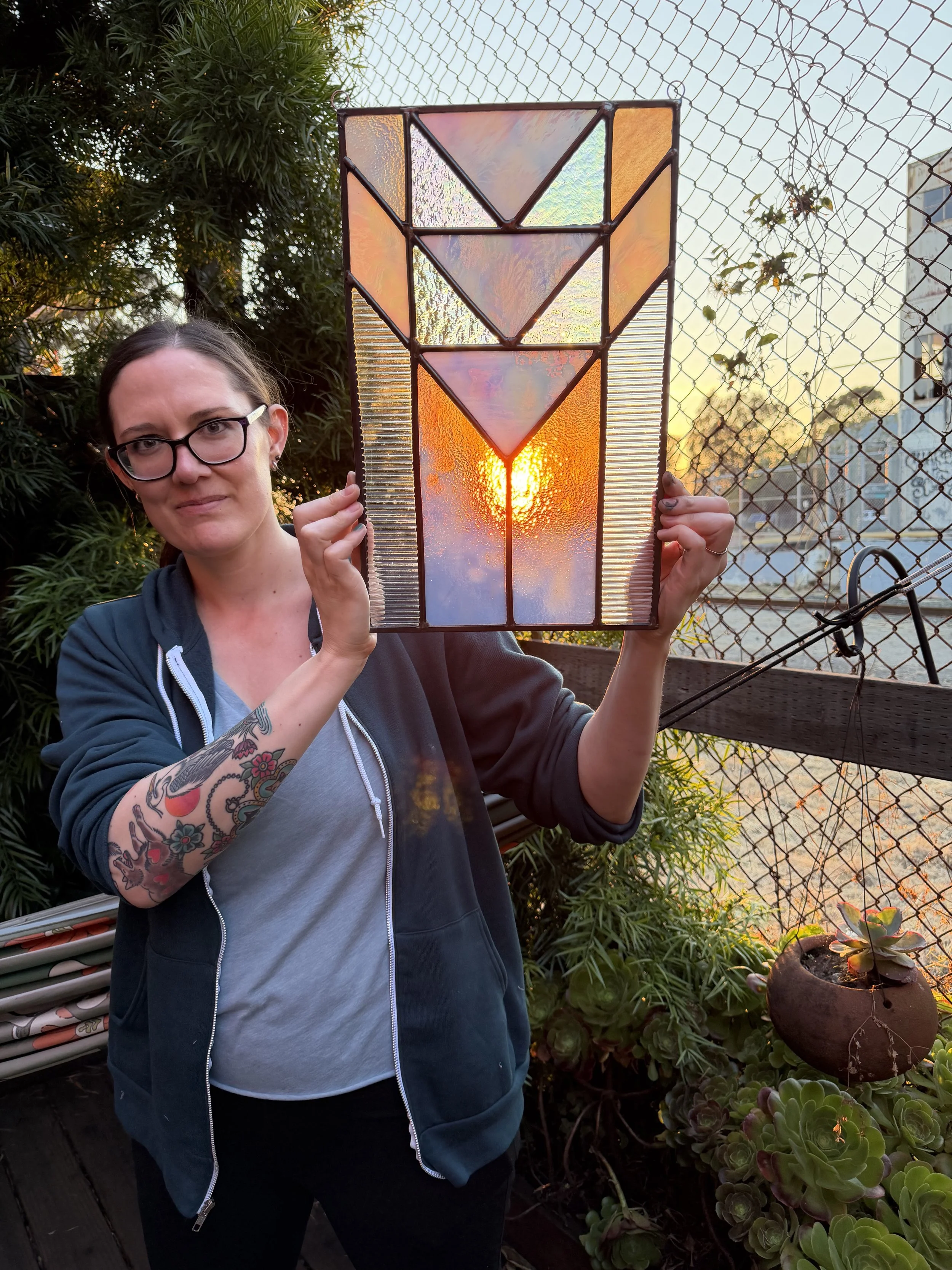 A woman with glasses and tattoos on her arm is holding a stained glass panel with a sunset or sunrise background, outdoors on a balcony with plants and a wire fence in the background.