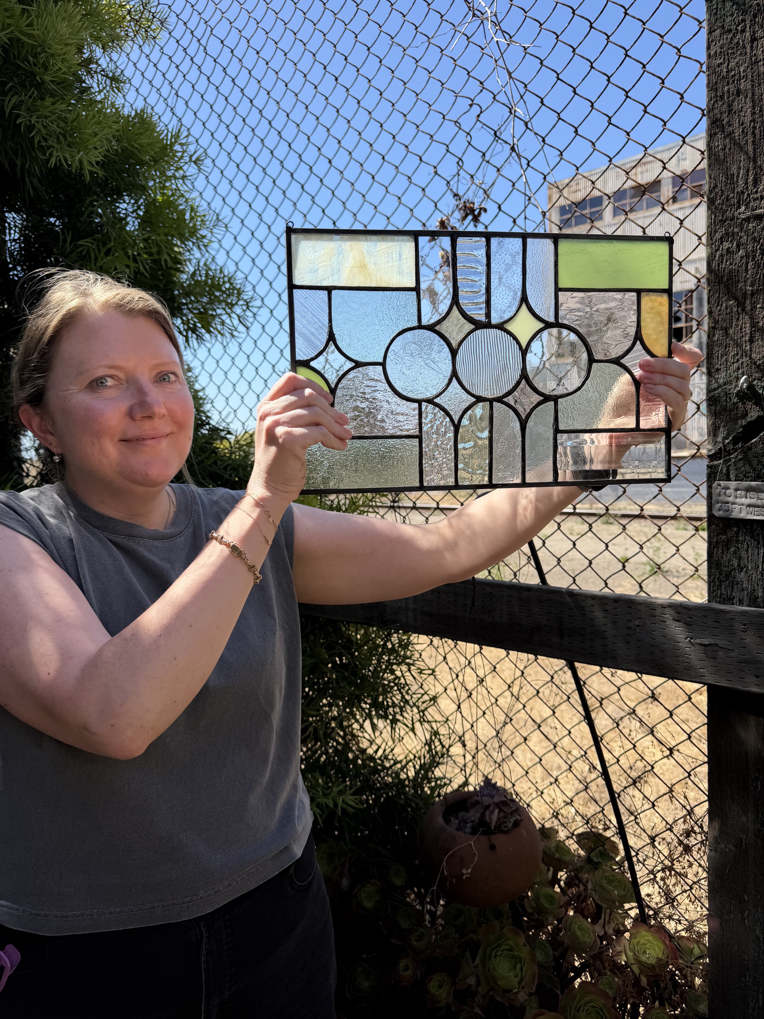 A woman holding a stained glass panel outdoors near a chain-link fence, with green plants and a building in the background.