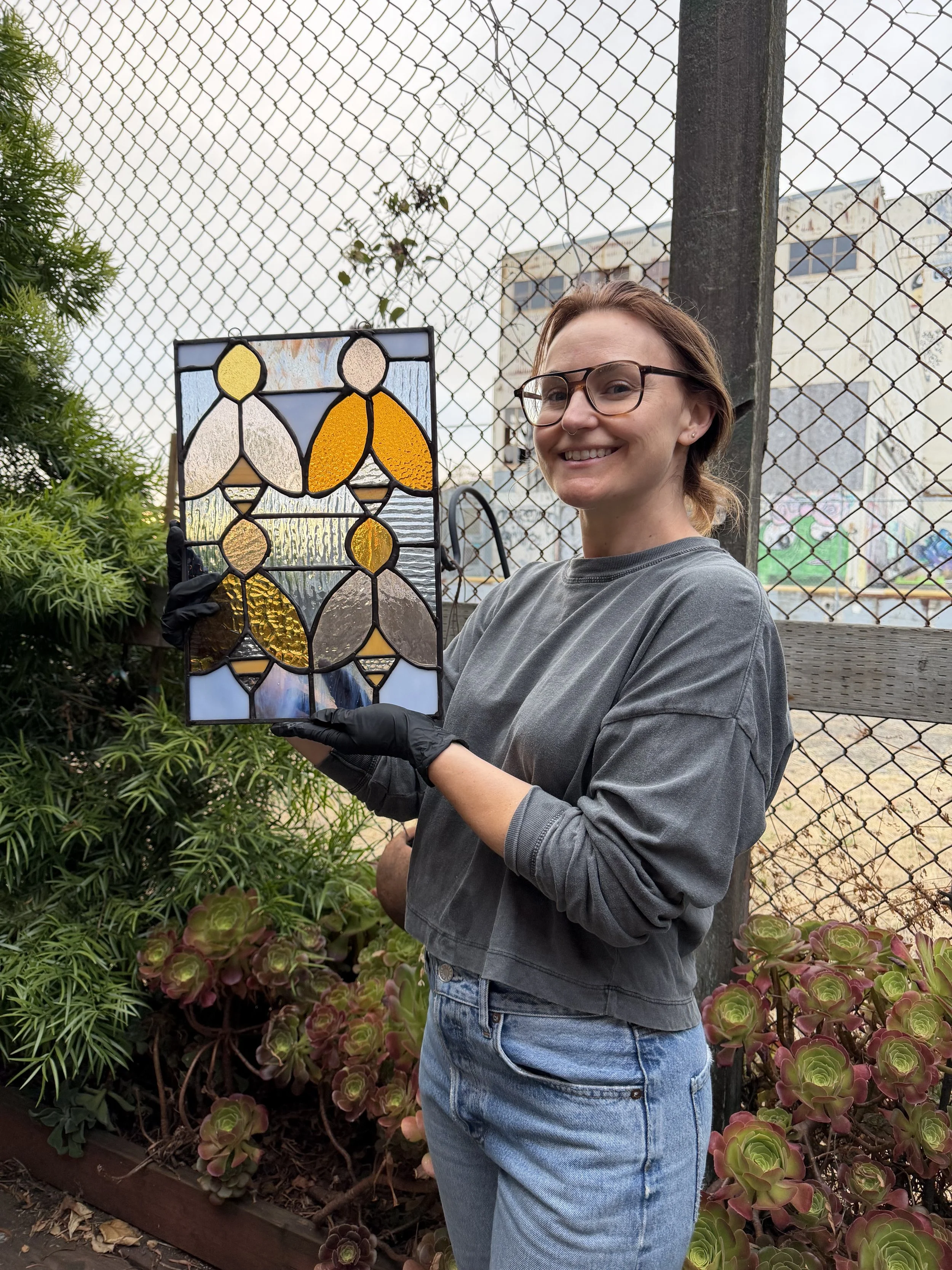 A woman with glasses, a gray shirt, and jeans holding a stained glass artwork outdoors in front of a chain-link fence with plants and buildings in the background.