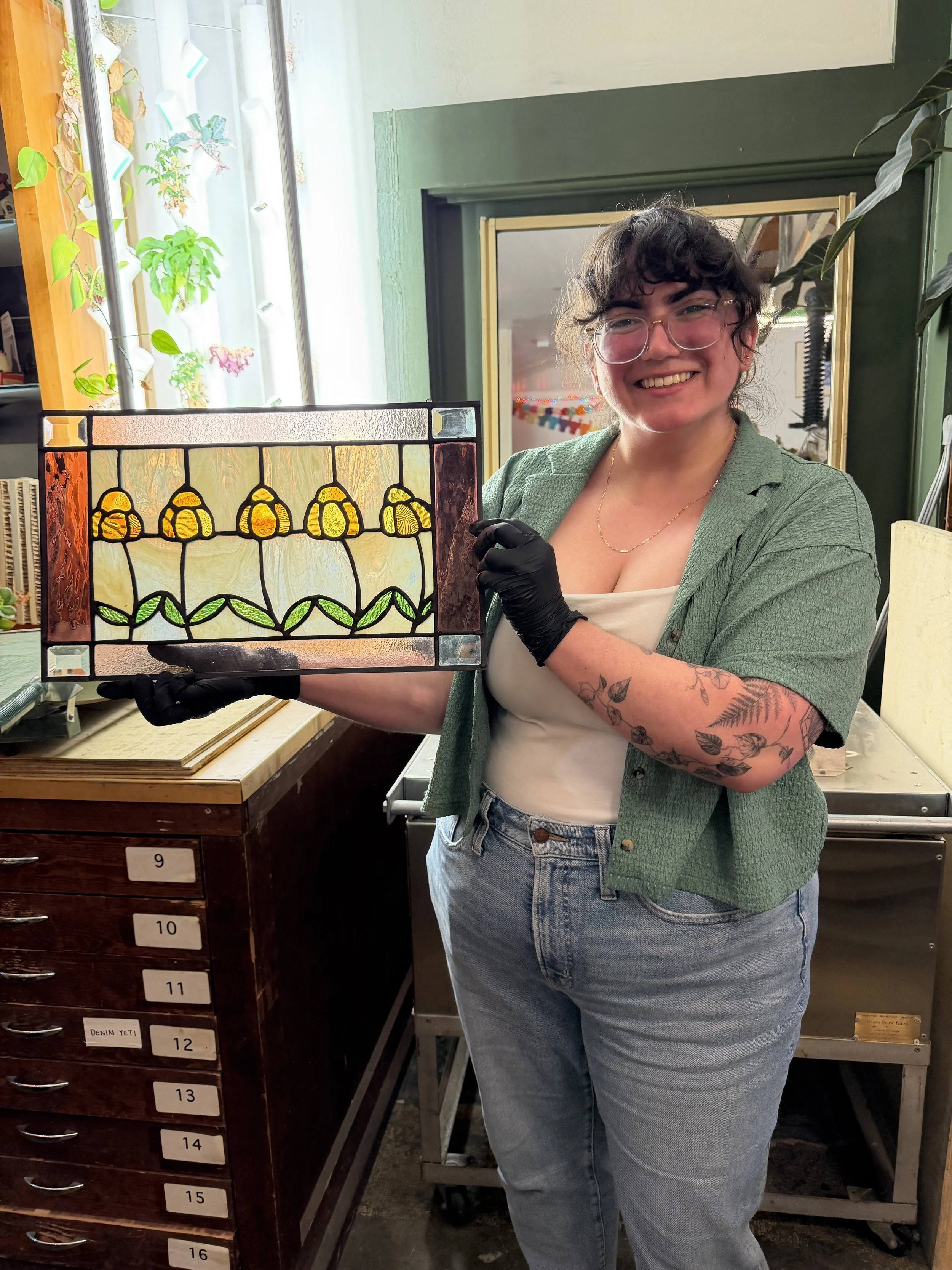 A woman with curly hair, glasses, and tattoos on her arm is smiling and holding a stained glass panel with a floral design in an art or craft studio.