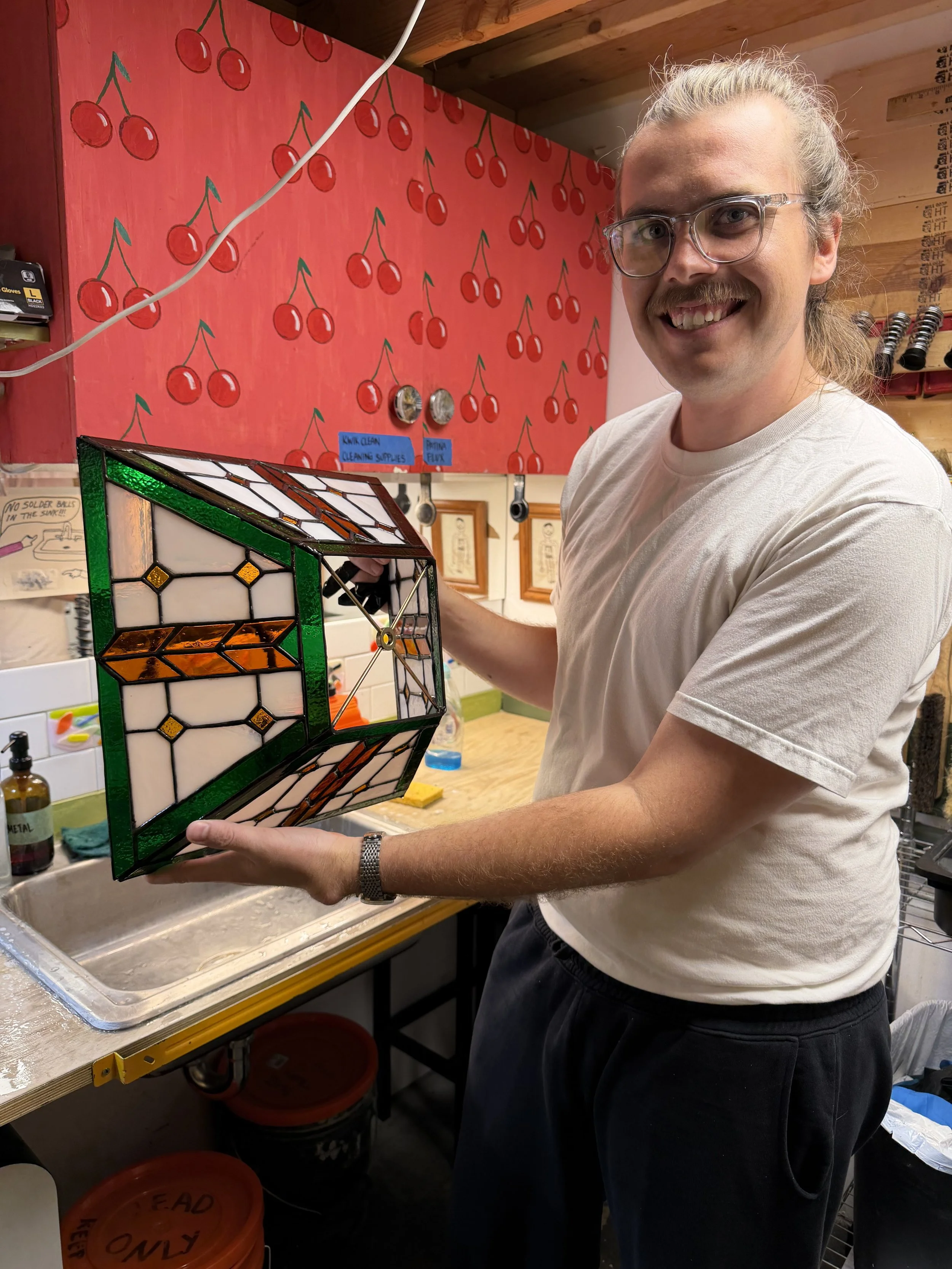 A man with glasses and a mustache smiling while holding a stained glass lamp in a workshop. There are cherry-patterned wall decorations and tools in the background.
