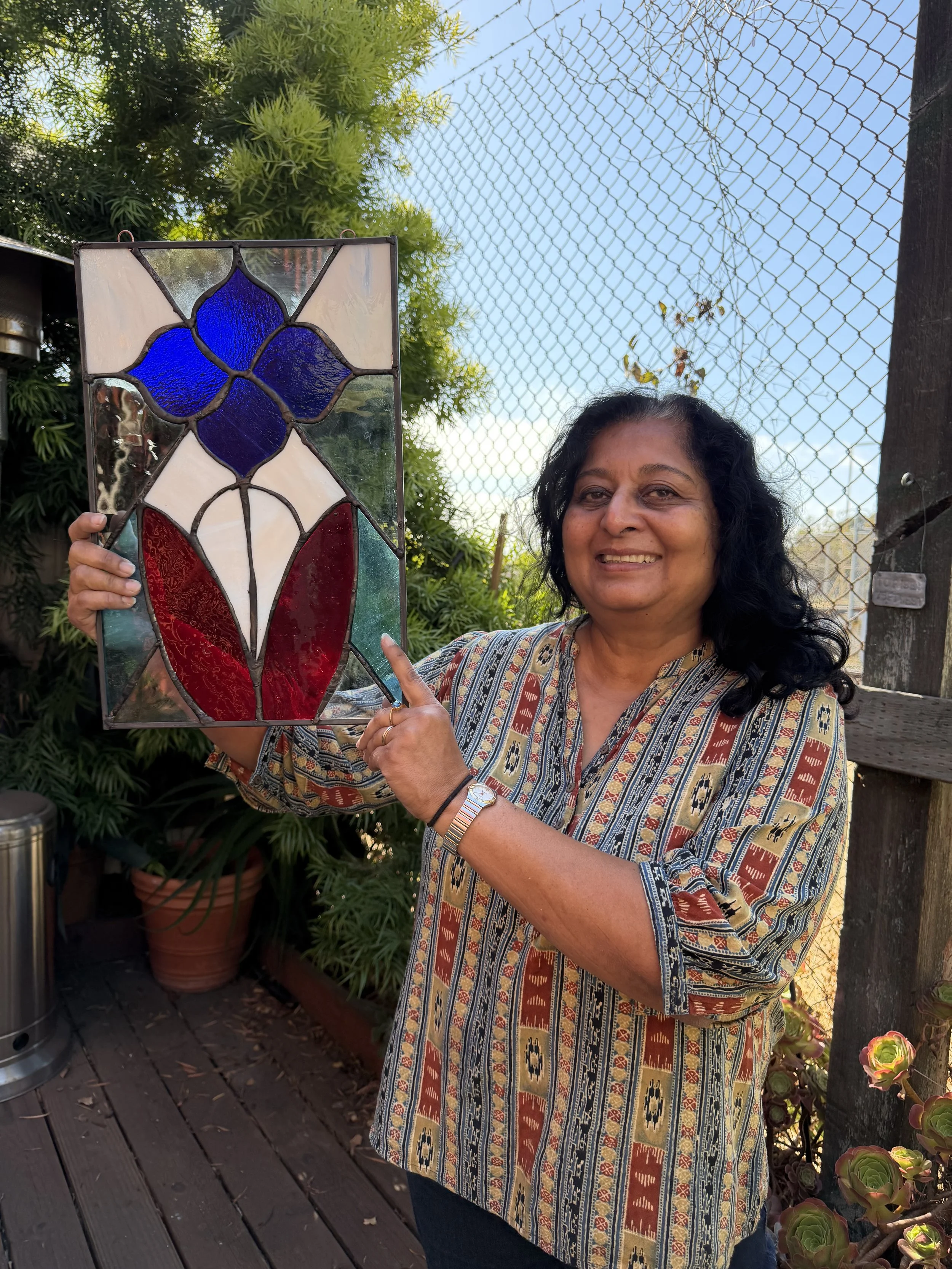 A woman smiling outdoors holding a stained glass artwork with a blue flower design, standing on a wooden deck with potted plants and a chain-link fence in the background.
