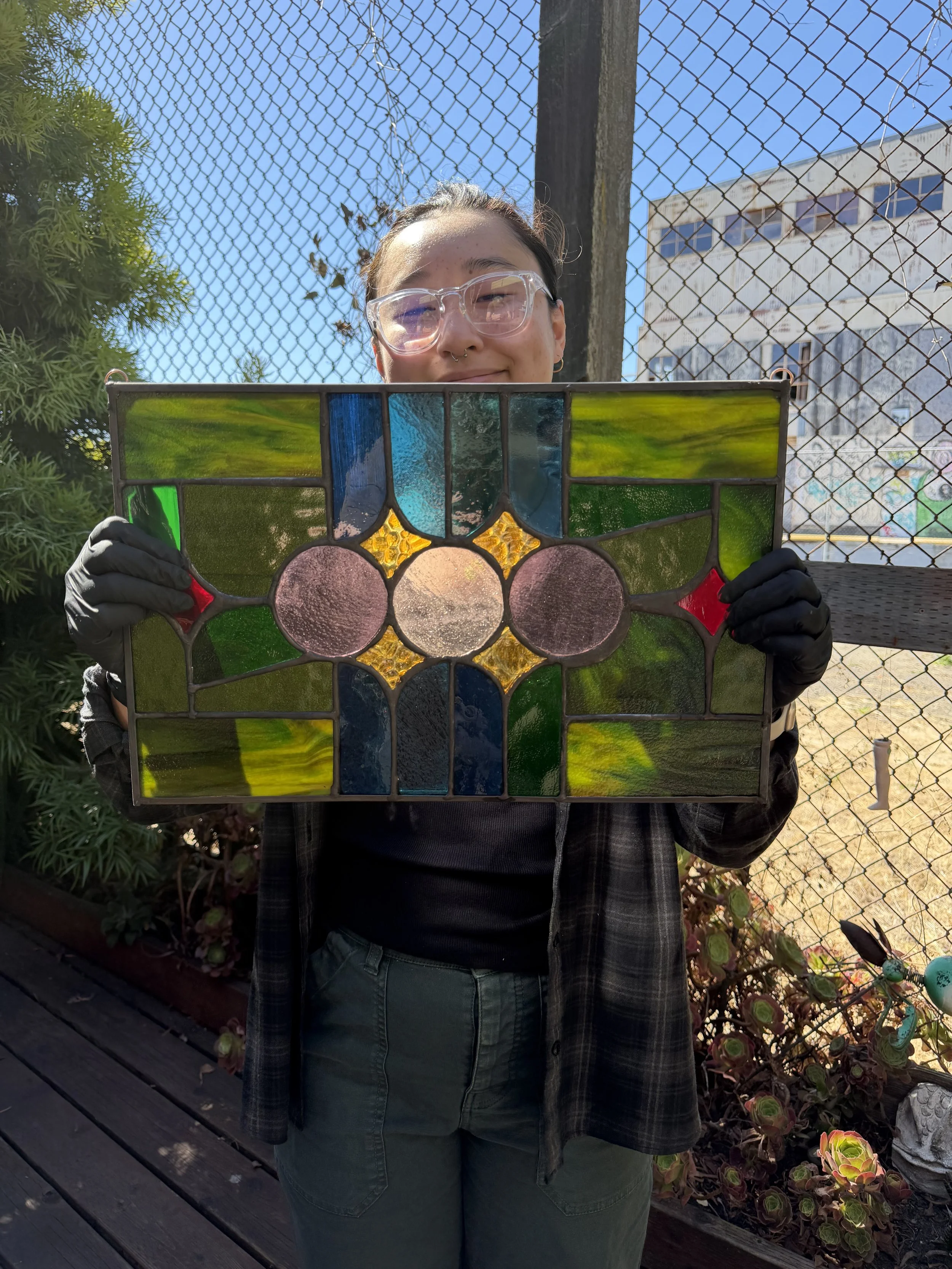 Person holding a stained glass panel outdoors, fencing and a building visible in the background.