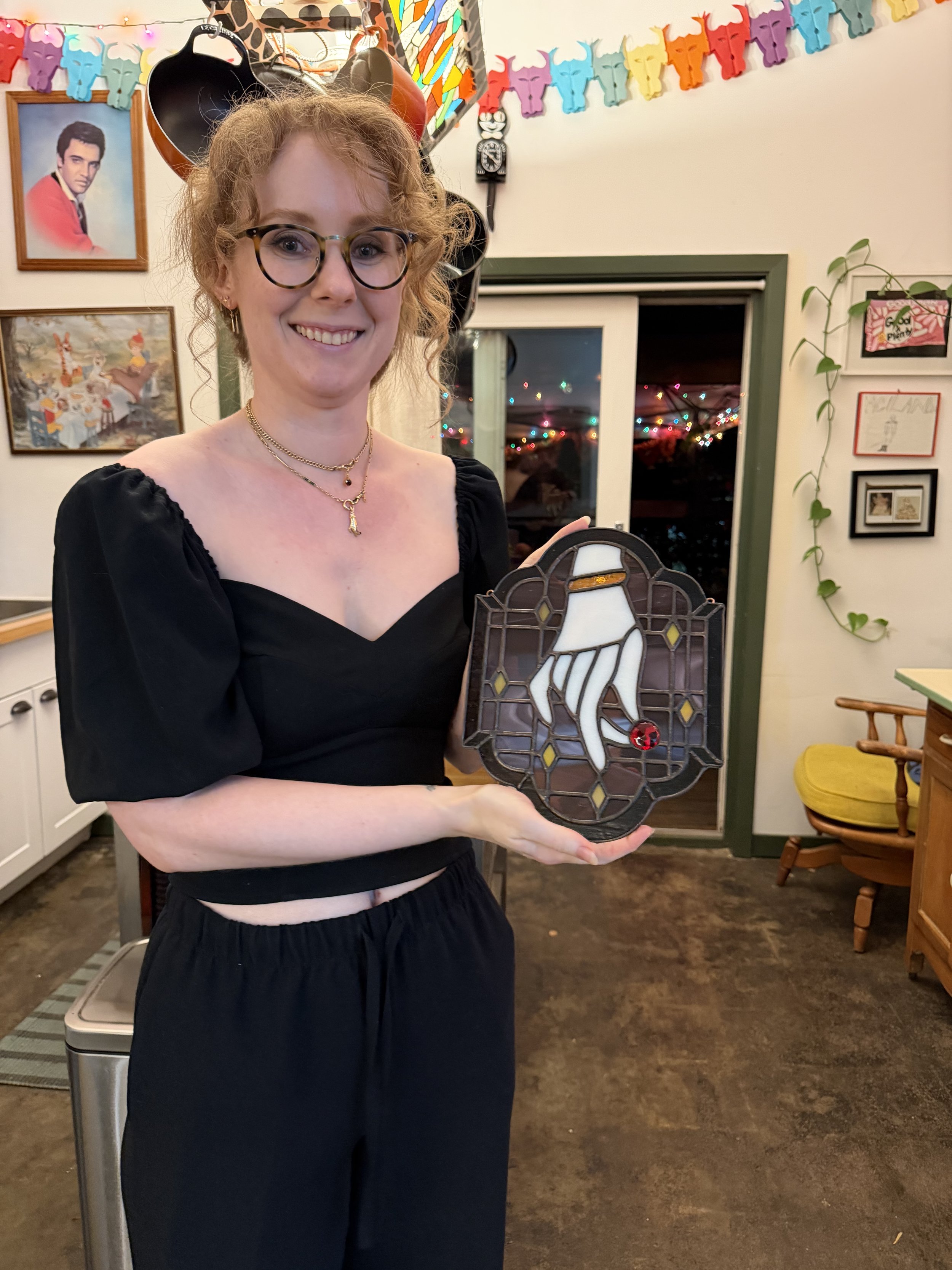 A woman with curly blonde hair and glasses smiling at the camera while holding a stained glass panel featuring a dove with an olive branch.