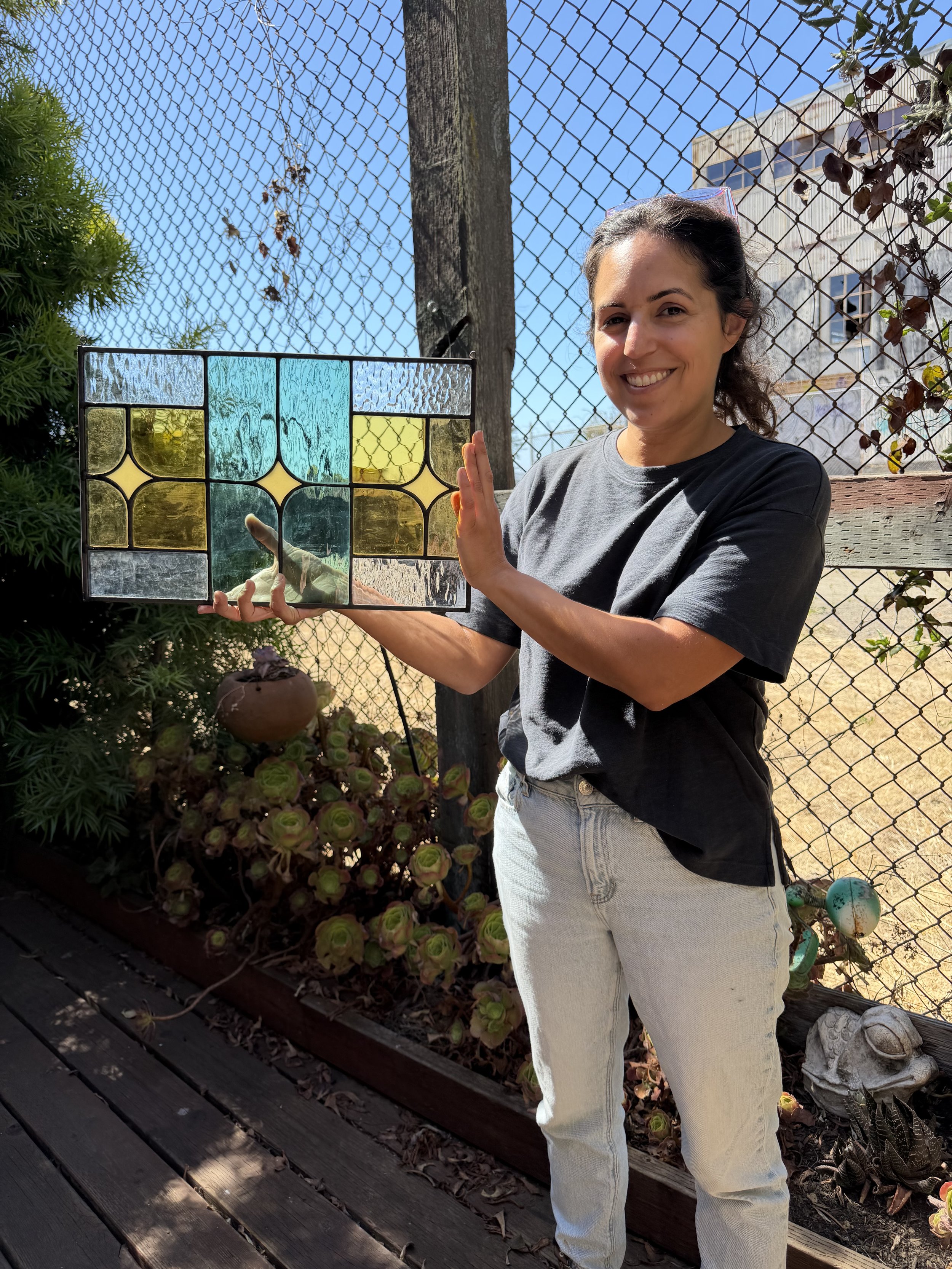 A woman smiling outdoors holding a stained glass window with colorful textured glass panels, standing in front of a chain-link fence and garden with succulent plants.