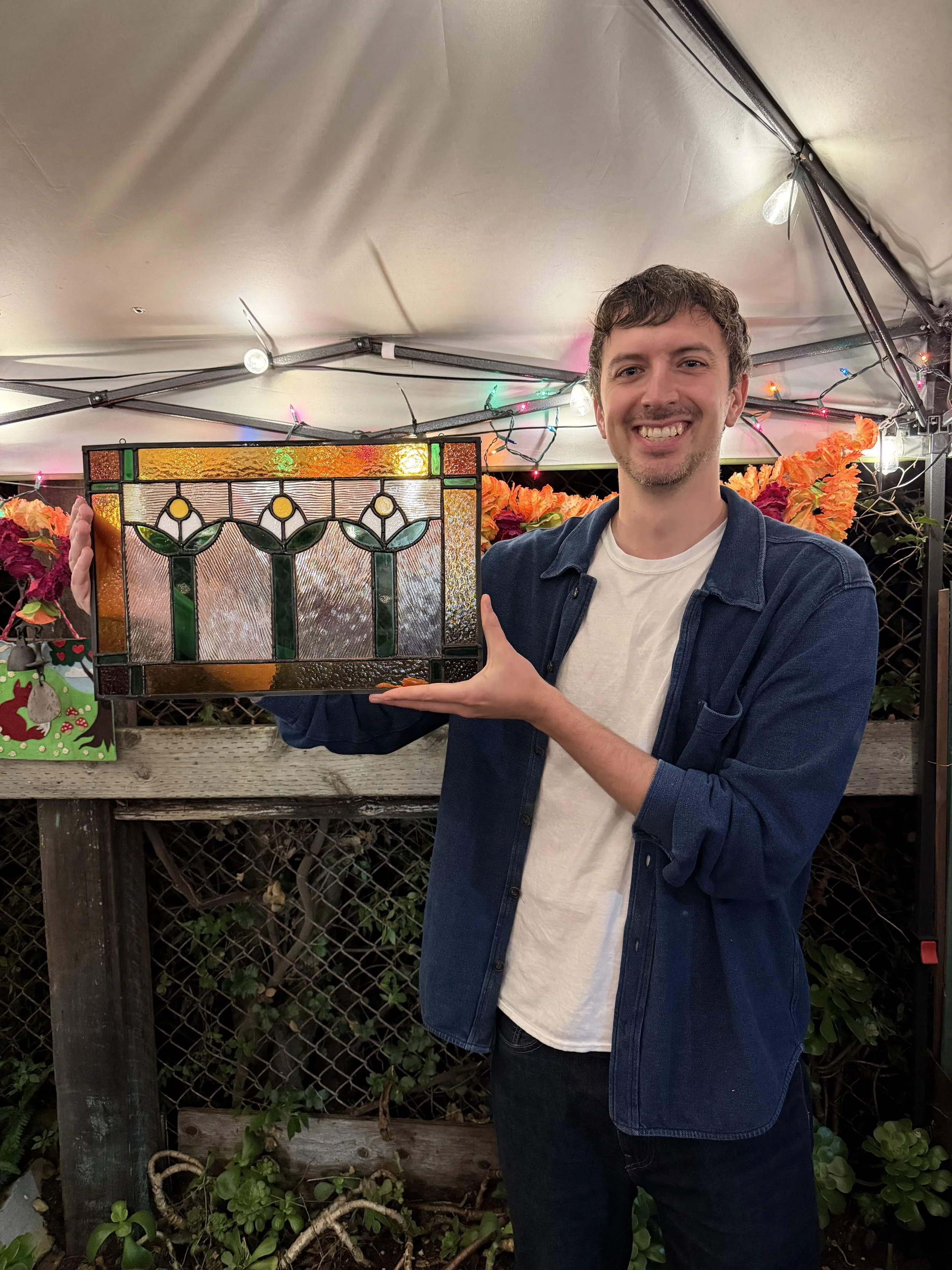 A smiling man holding a colorful stained glass artwork shaped like tulips in an outdoor setting under a canopy with string lights.