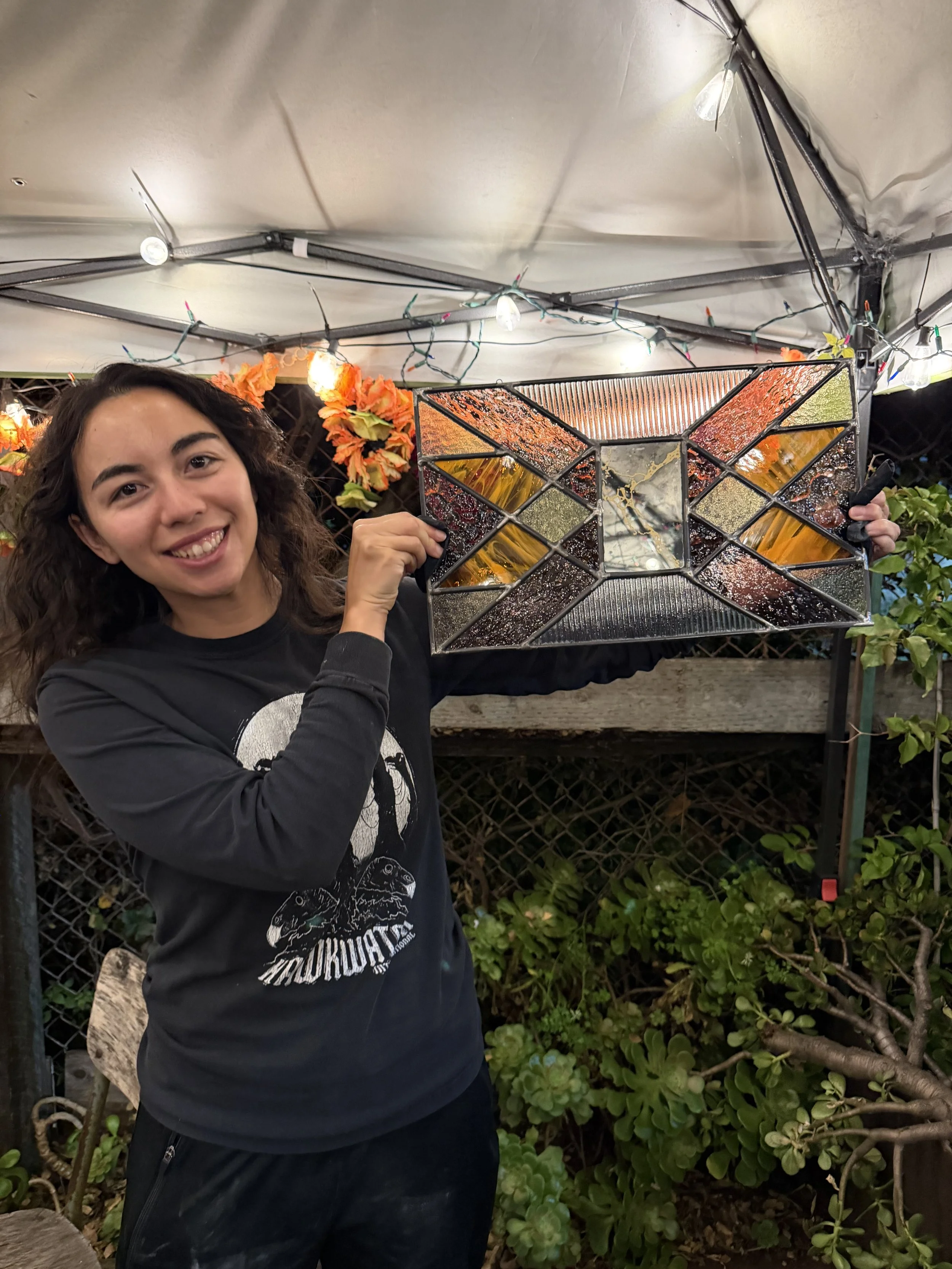 Young woman smiling and holding a decorative stained glass mirror in an outdoor garden setting at night, with string lights and flowers overhead.