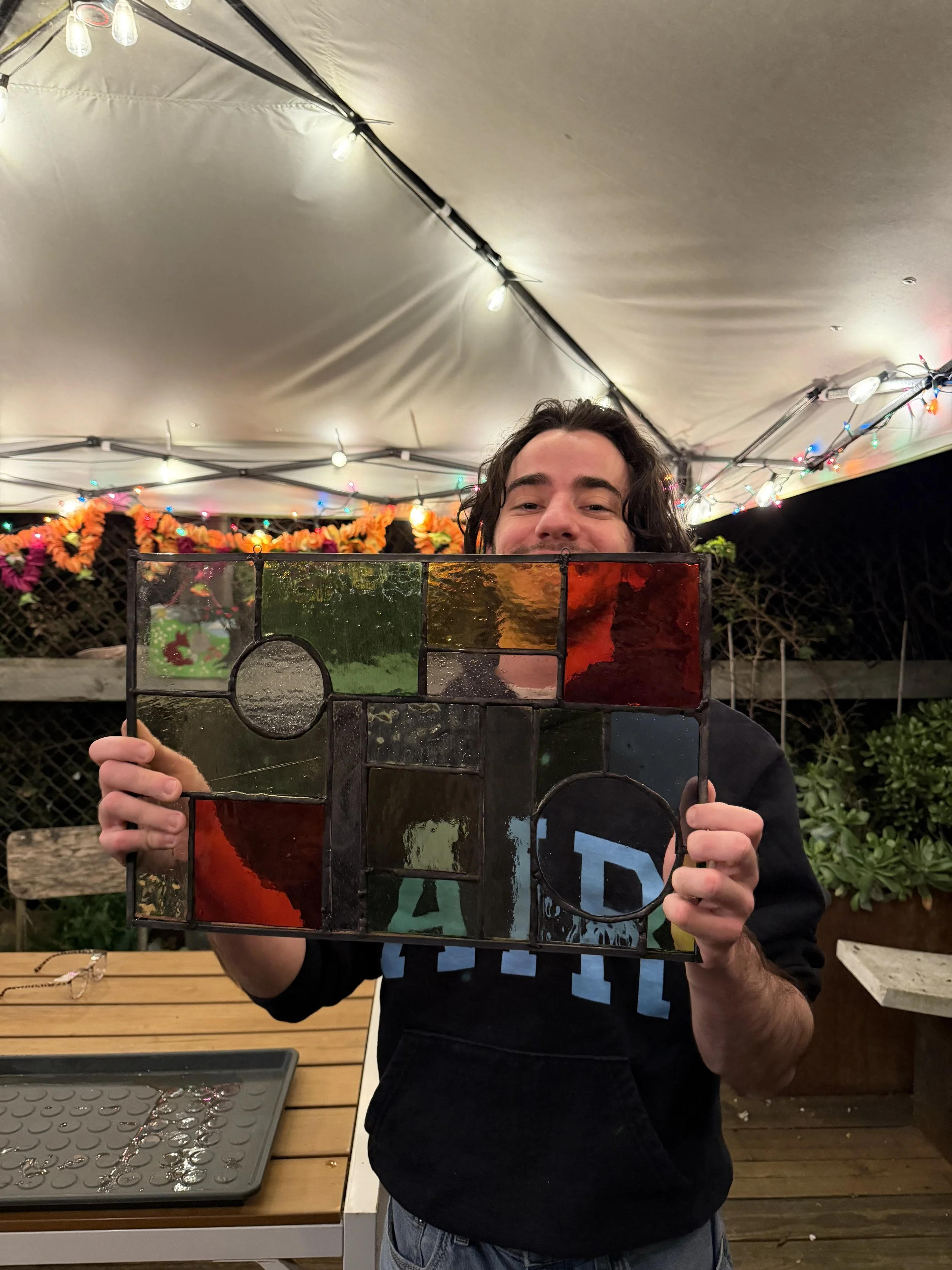 A man holding a colorful stained glass window panel with a festive background of string lights and flowers at night.