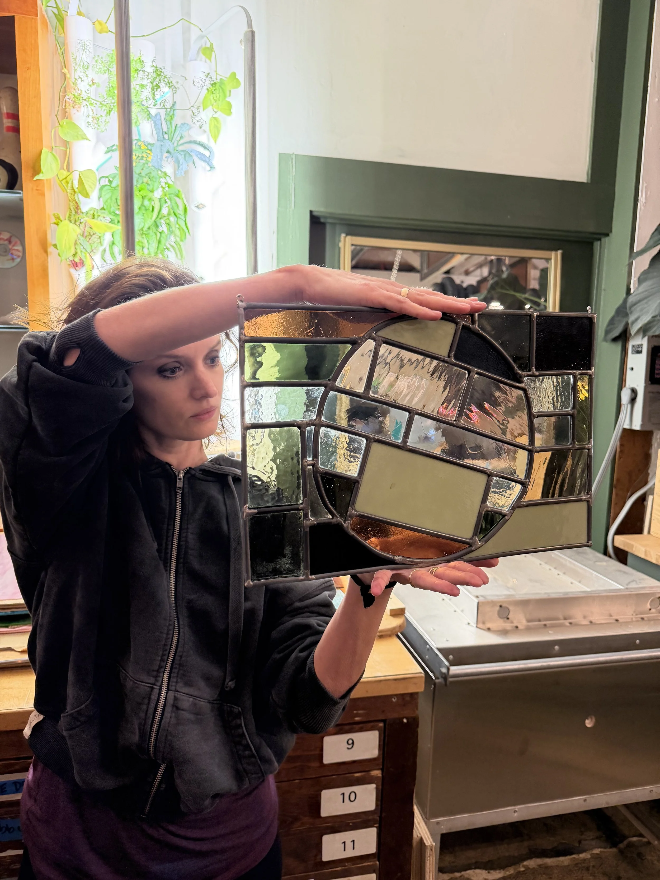 A woman is examining a stained glass piece in a workshop, holding it up to her face. The stained glass has various shapes and colors, with a focus on earth tones and geometric patterns.
