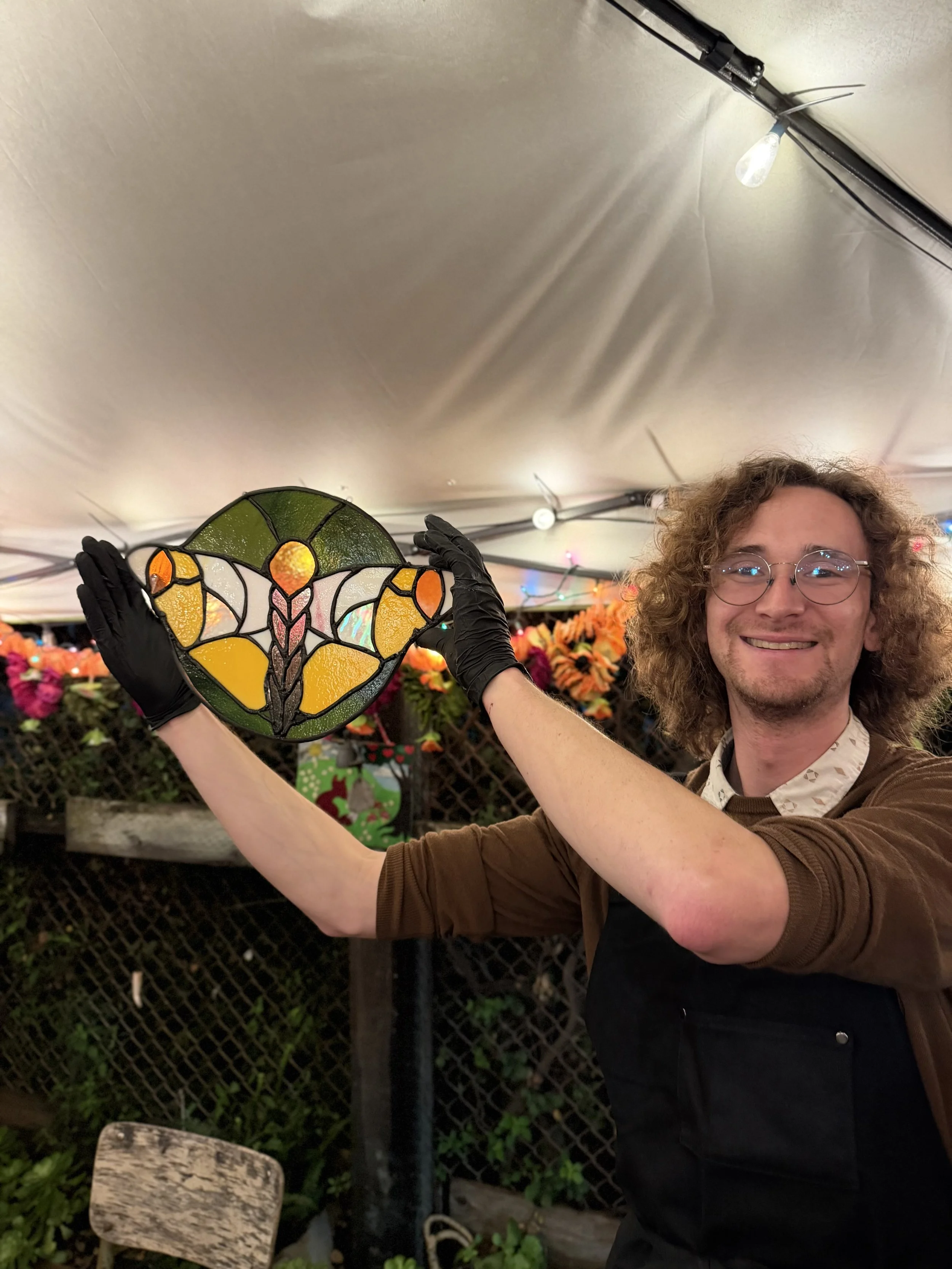 A person with curly hair and glasses holding a stained glass butterfly decoration at an outdoor event with tables of flowers and string lights in the background.