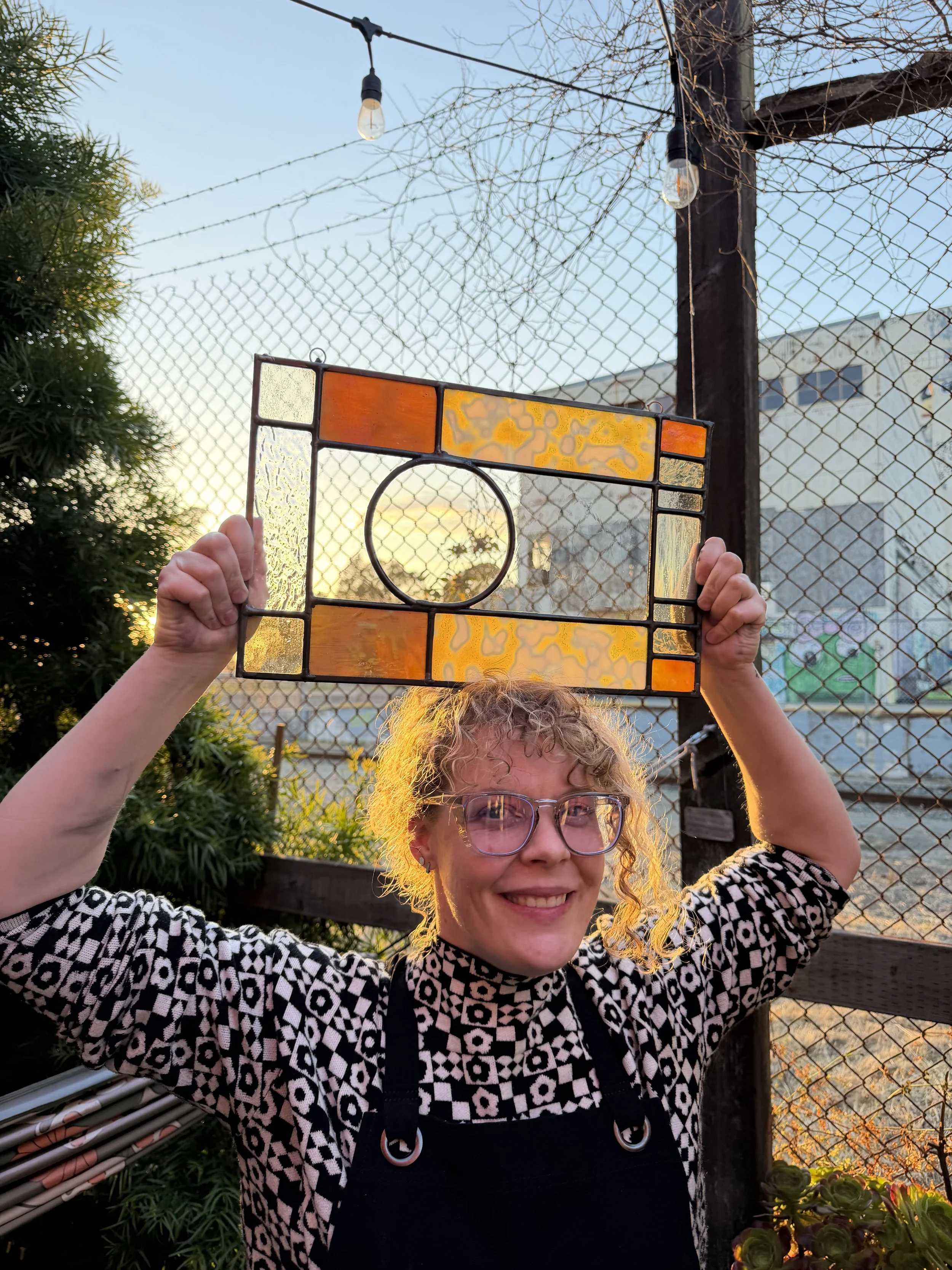 A woman with curly blonde hair and glasses is smiling outdoors during sunset, holding a stained glass window with orange, yellow, and clear glass pieces above her head. She is standing in front of a chain-link fence and some greenery.