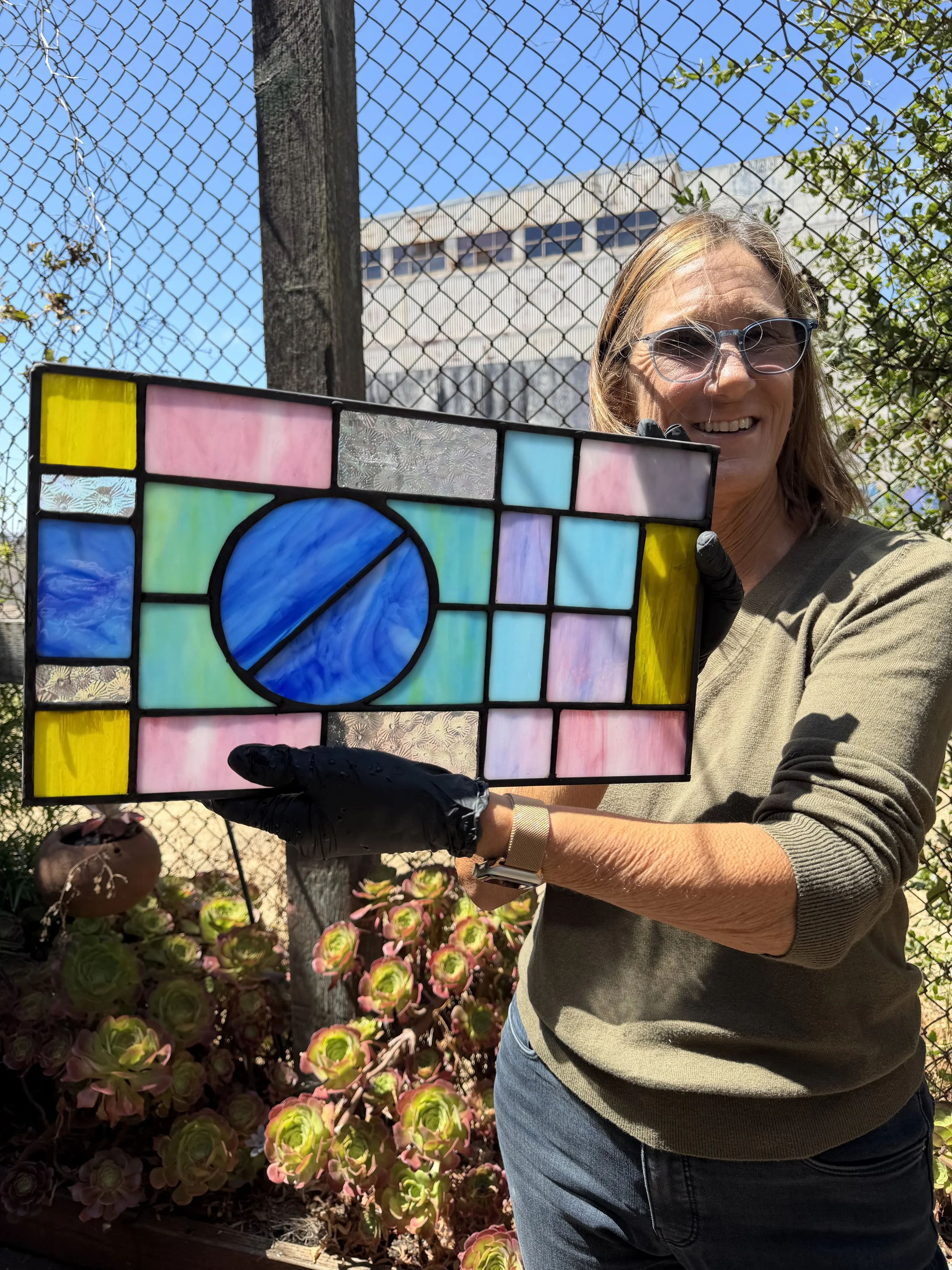 A woman smiling, holding a colorful stained glass piece in front of a garden, with a chain-link fence and a building in the background.
