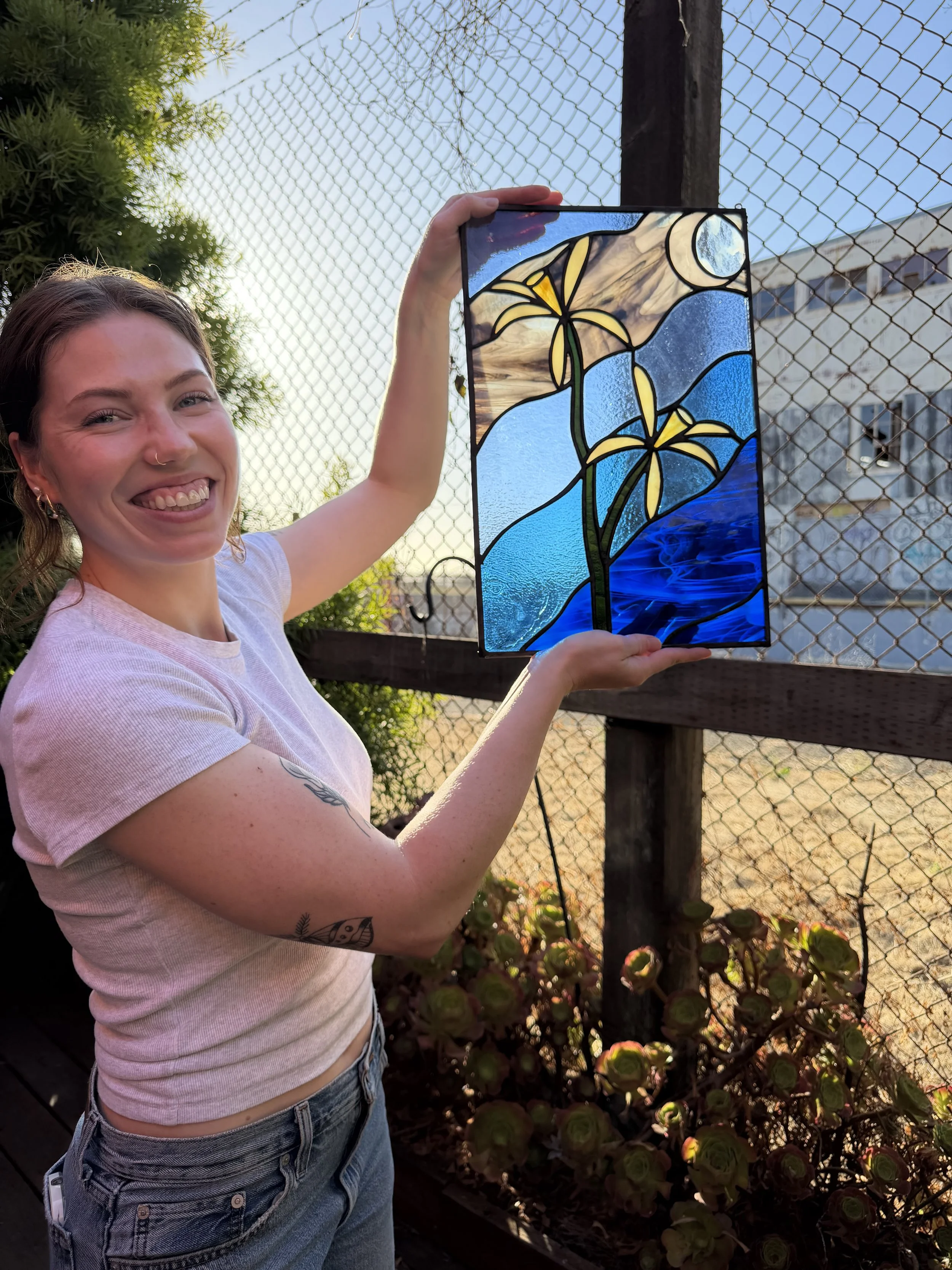 A young woman smiling and holding up a stained glass artwork featuring yellow flowers, blue water, and a moon, outdoors near a fence and greenery.
