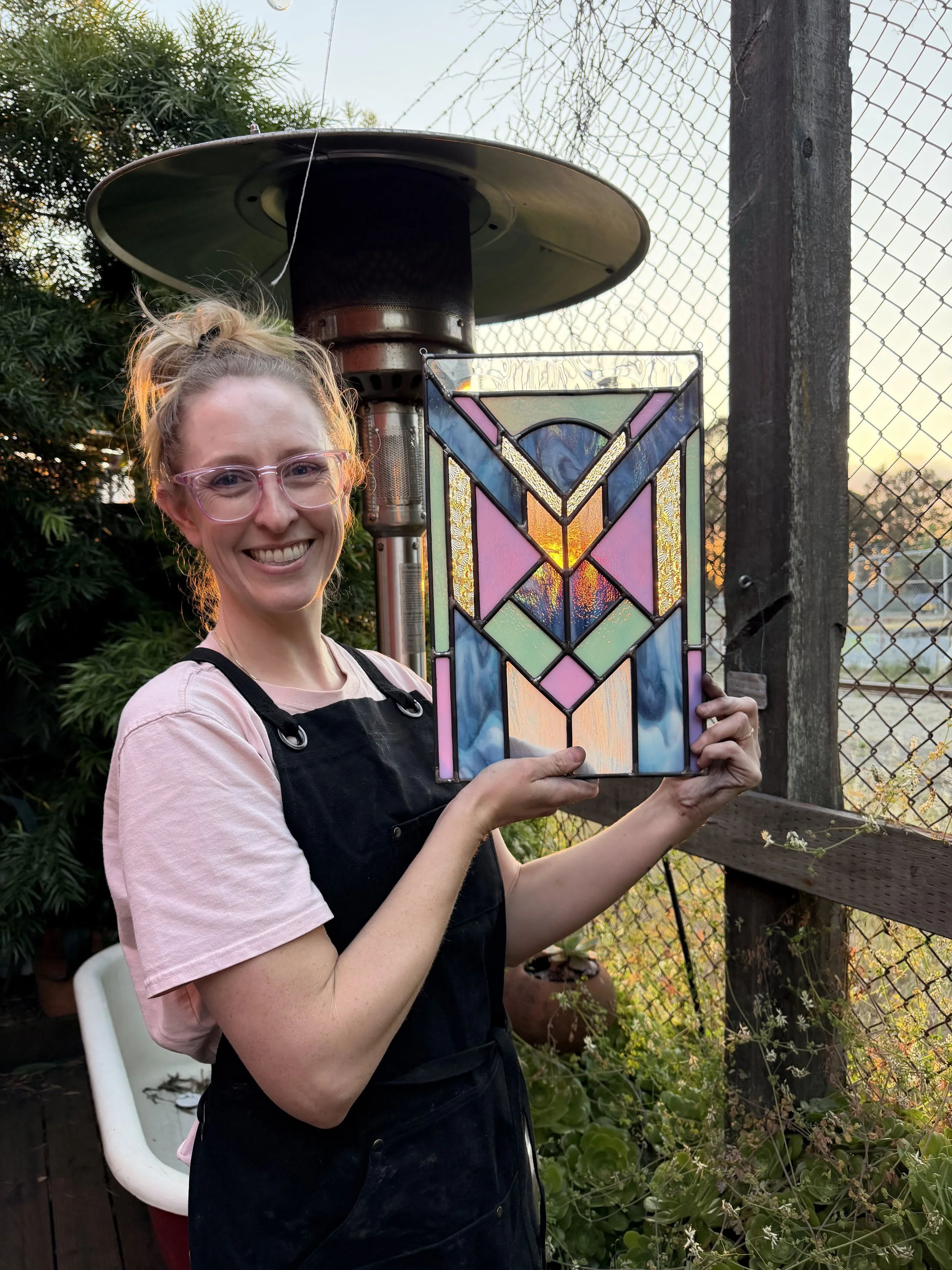 A smiling woman with pink glasses holding a colorful stained glass art piece outdoors during sunset. She is wearing a pink t-shirt and a black apron, with a garden and outdoor heater in the background.
