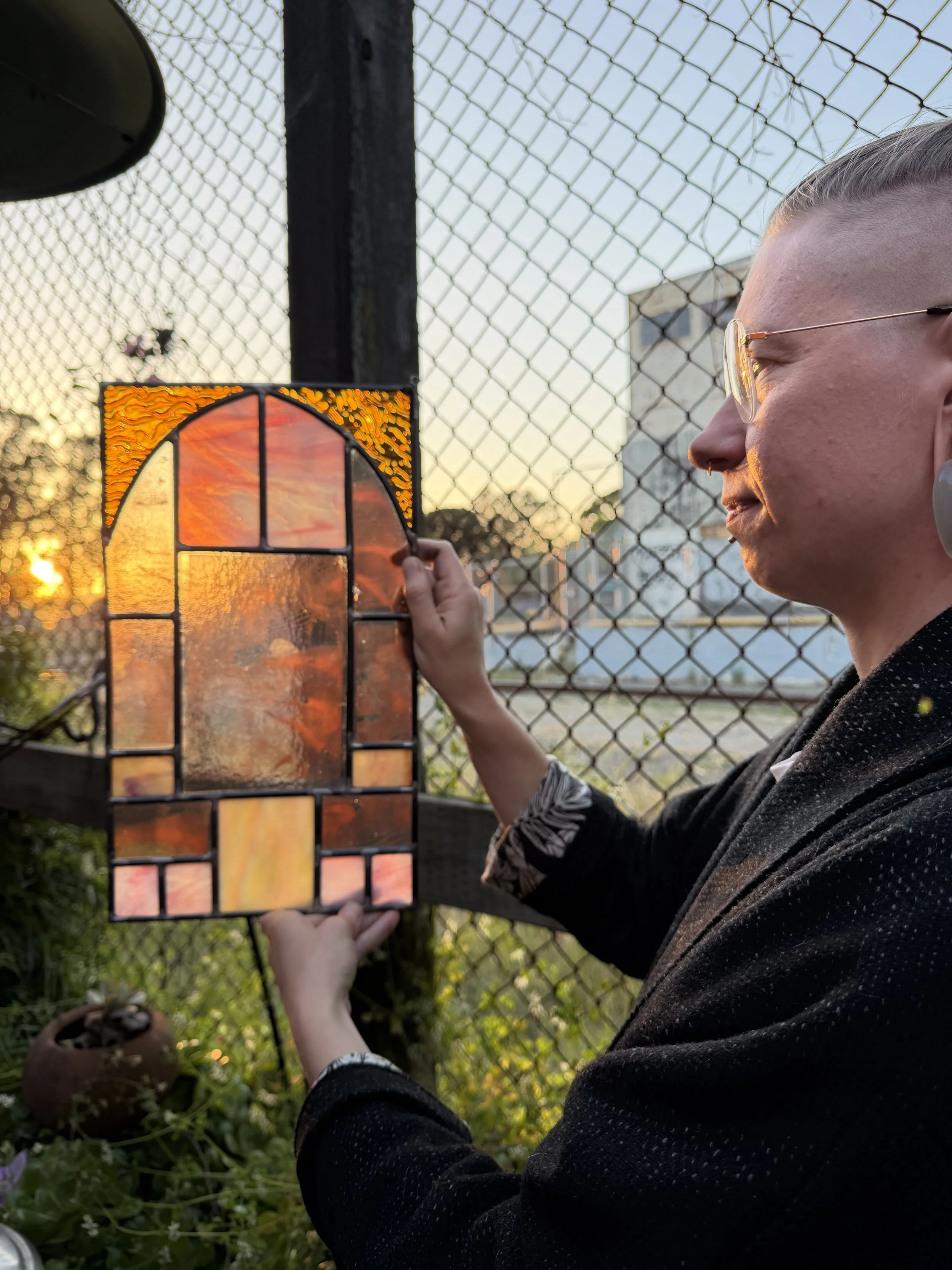 A person holding a piece of stained glass art in front of a sunset, with a chain-link fence and a building in the background.