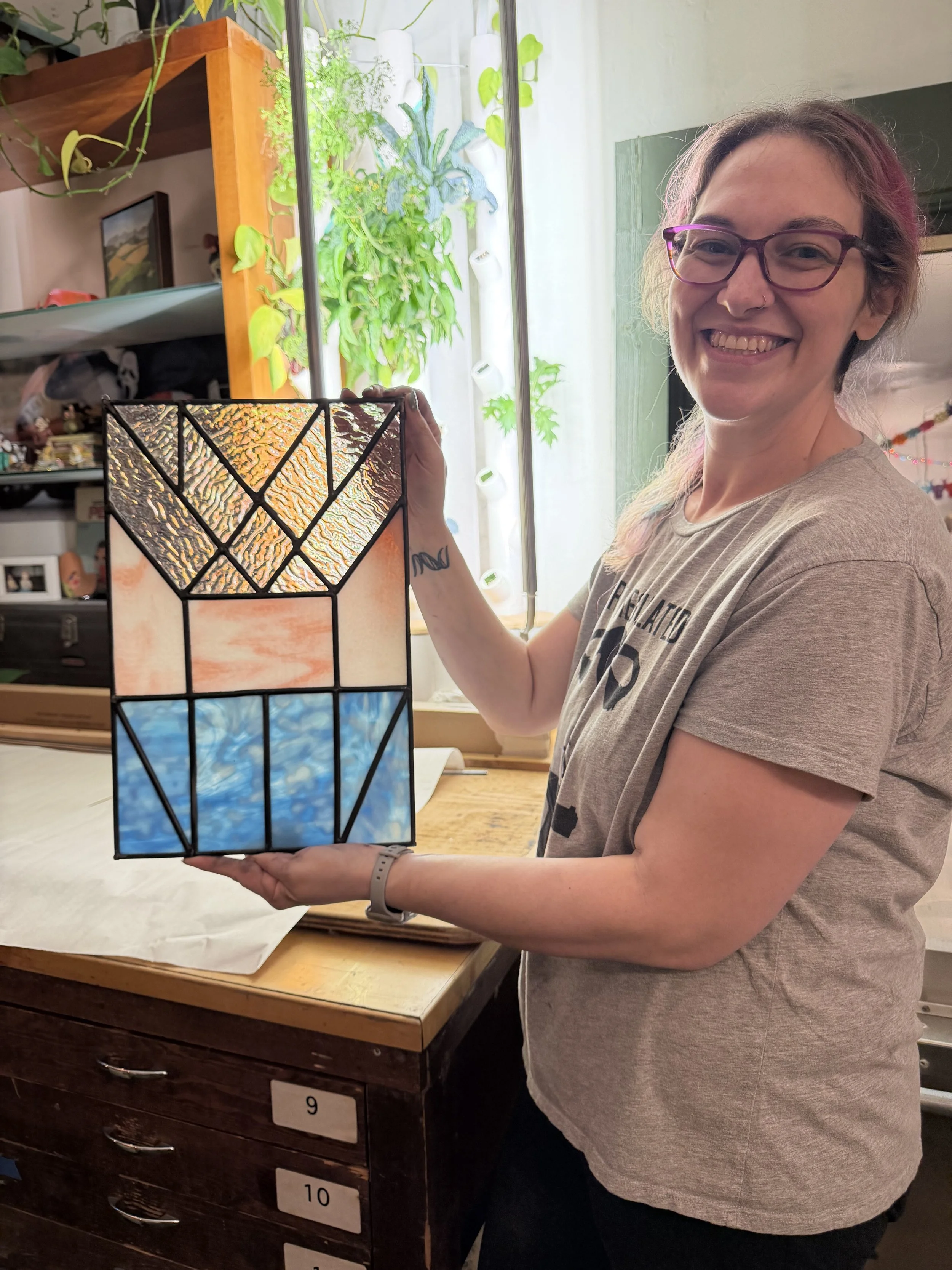 A woman with glasses and pink highlights in her hair smiling and holding a stained glass lamp with geometric patterns and colorful sections. She is standing indoors near a wooden storage unit with labeled drawers and a table covered with paper.