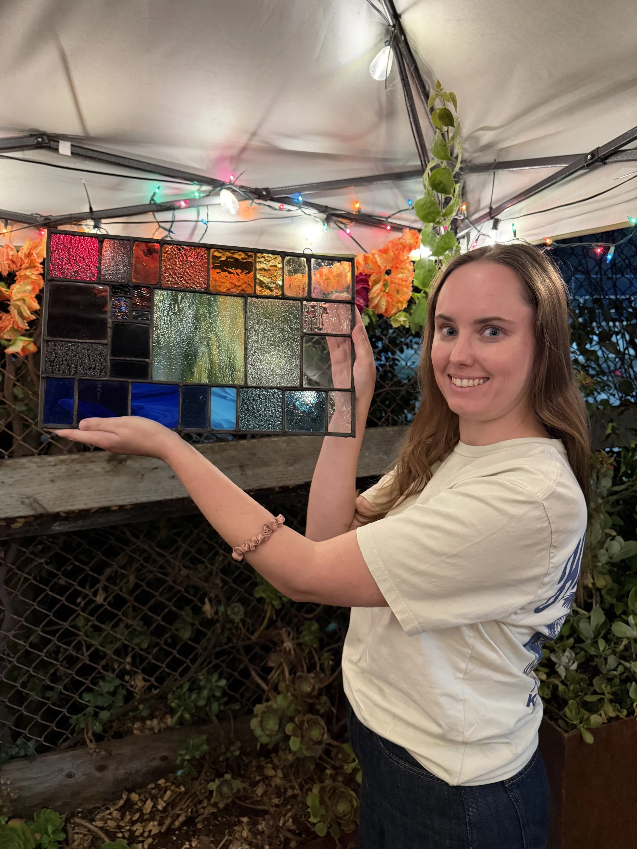 A young woman with brown hair smiling while holding a colorful stained glass panel outdoors under a canopy decorated with string lights, near plants and flowers.