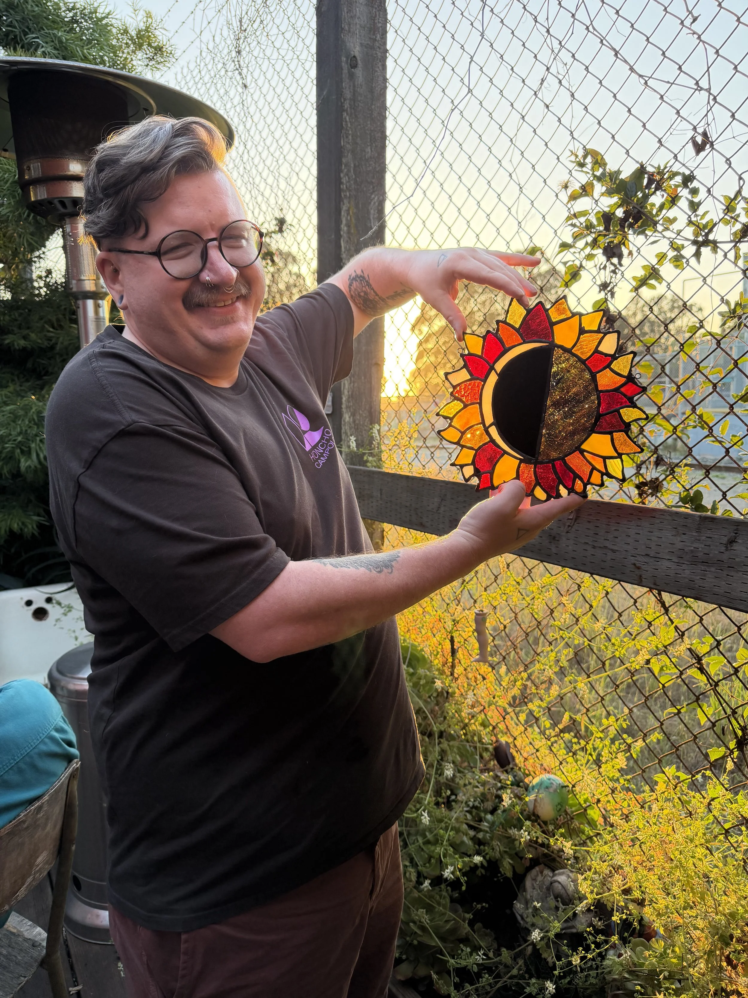 A person with glasses and tattoos on their arms holding a stained glass sun and moon ornament outdoors during sunset, with a chain-link fence and lush plants in the background.