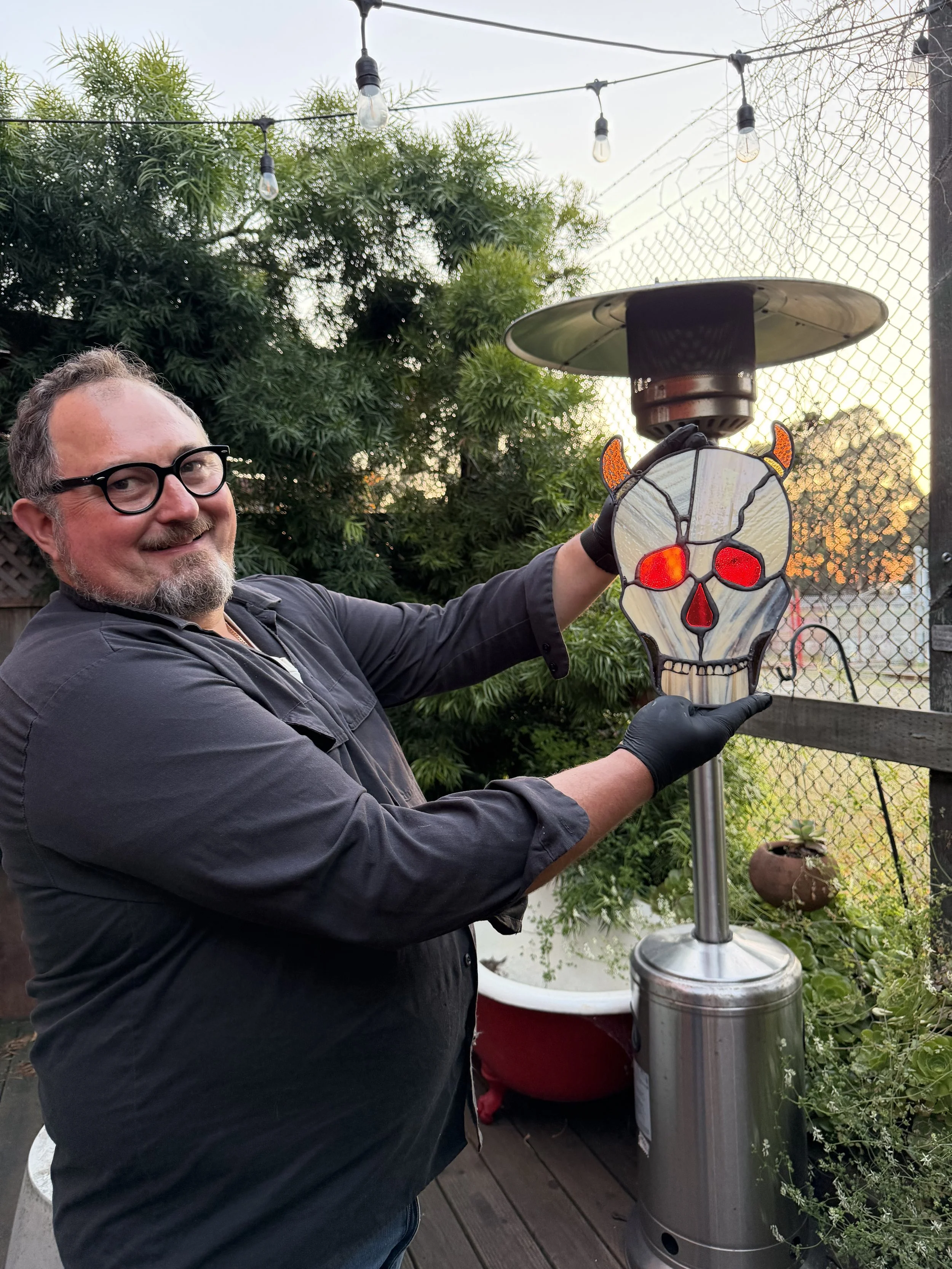 A man with glasses and a beard is smiling and holding a skull-shaped, stained glass decoration near a patio heater outdoors, with string lights and greenery in the background.