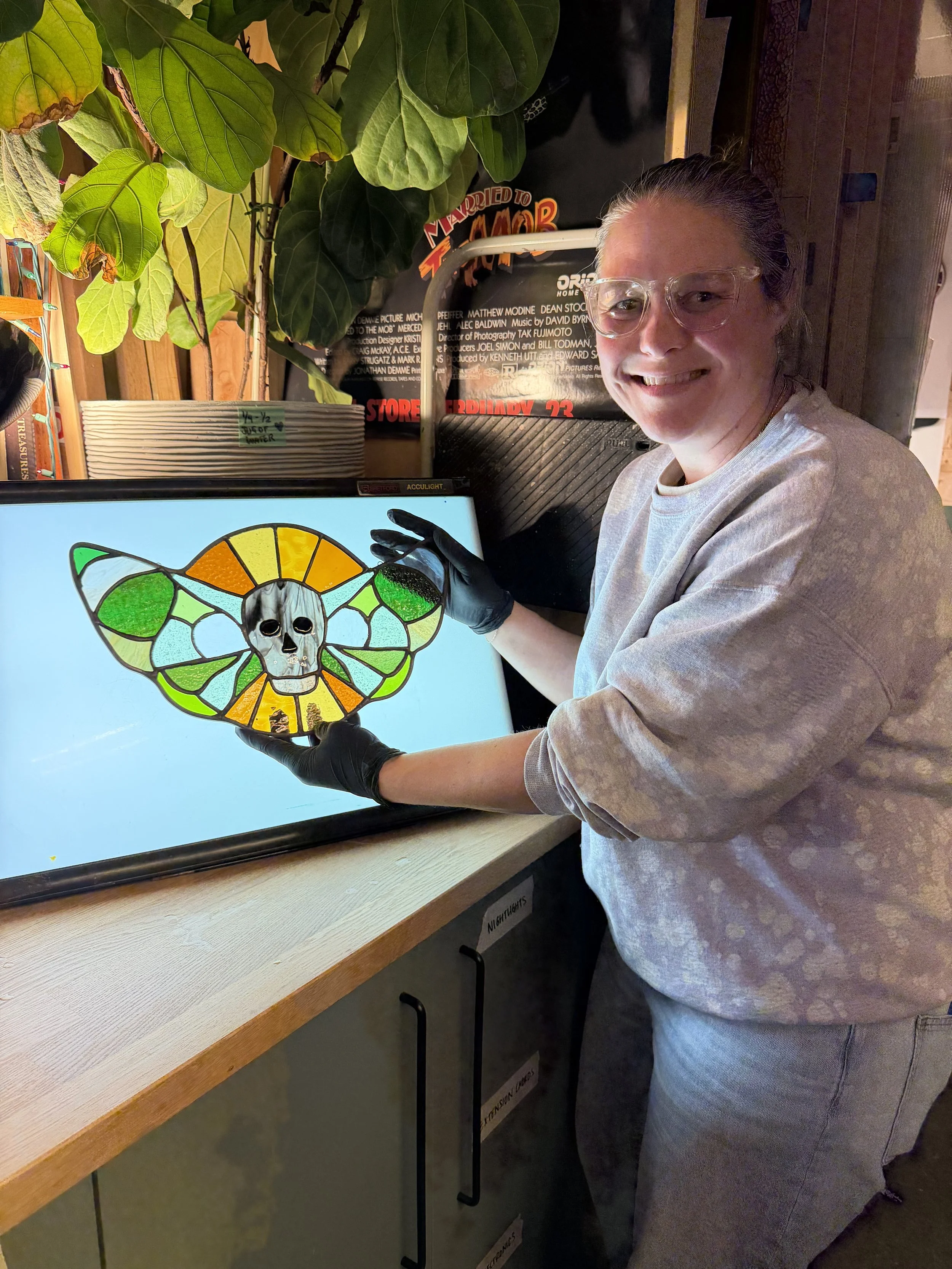 A woman with glasses and gray shirt smiling while holding a colorful stained glass art piece resembling a butterfly with a skull in the center.