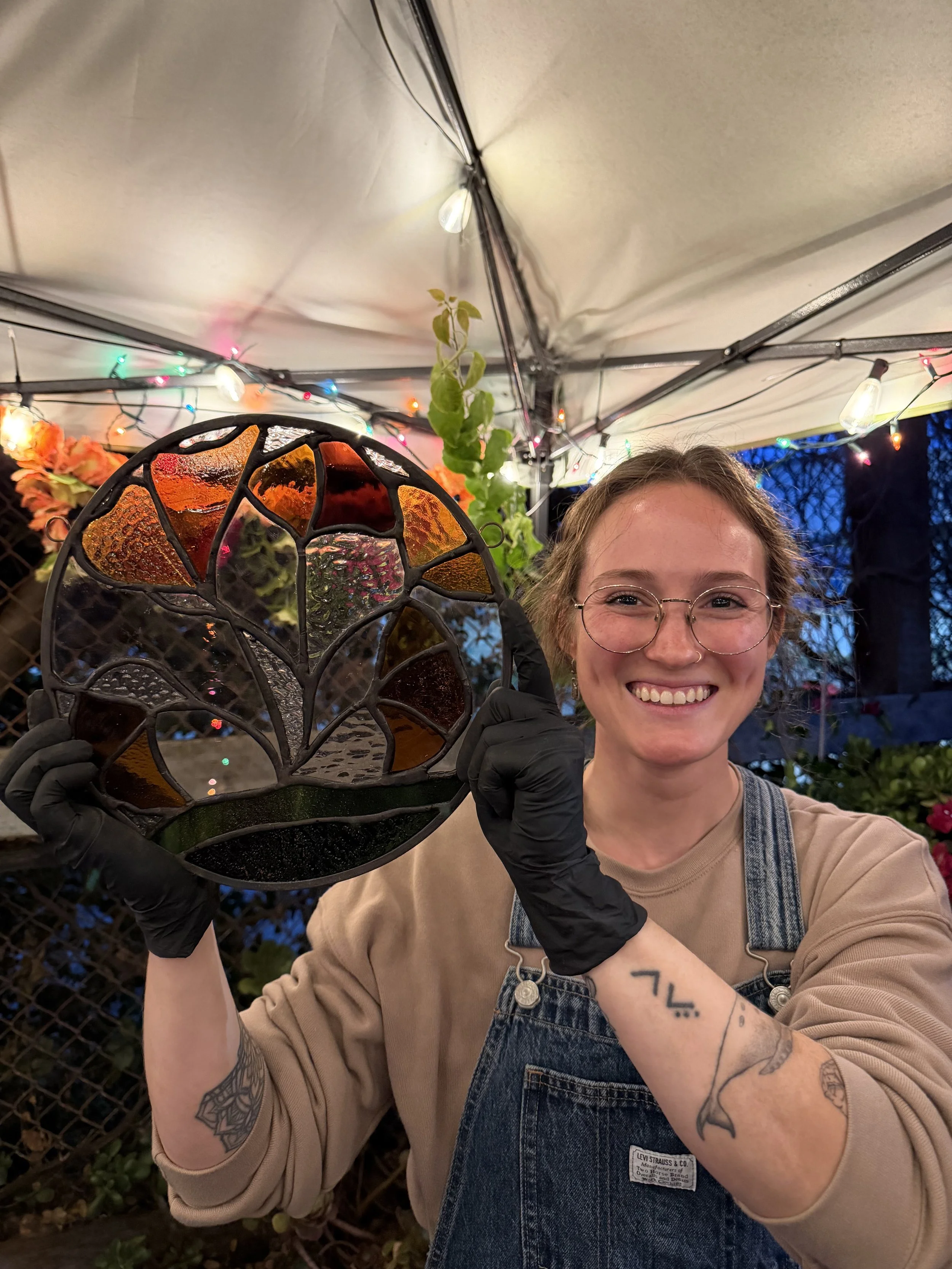 A smiling woman with glasses holding a stained glass ornament outdoors under a canopy decorated with string lights and flowers.