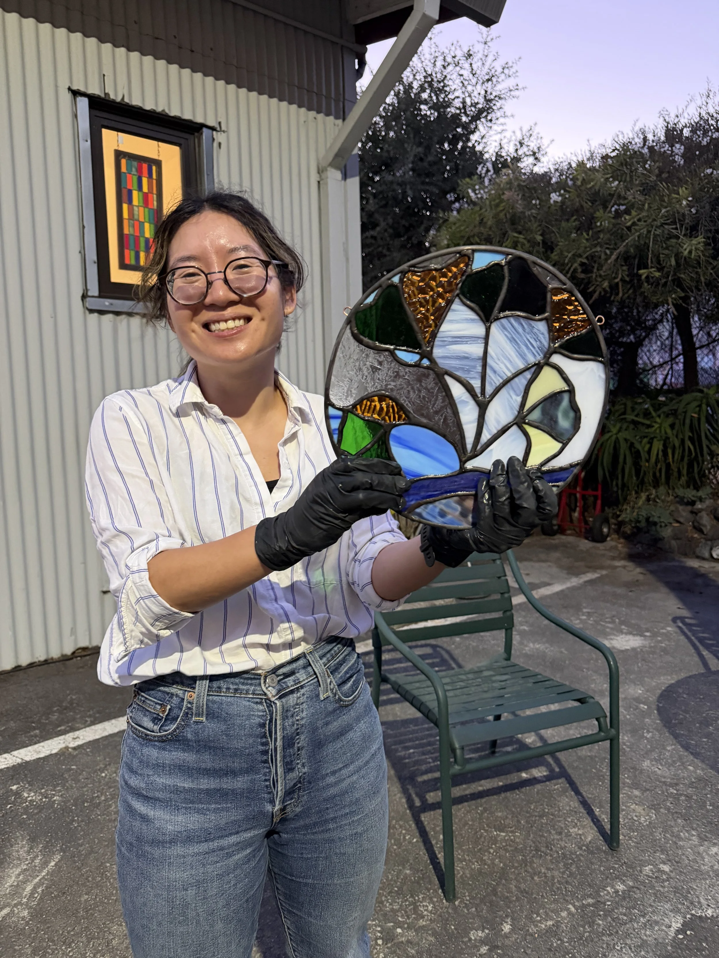 A woman with glasses and a striped shirt smiling and holding a stained glass art piece outdoors during dusk.