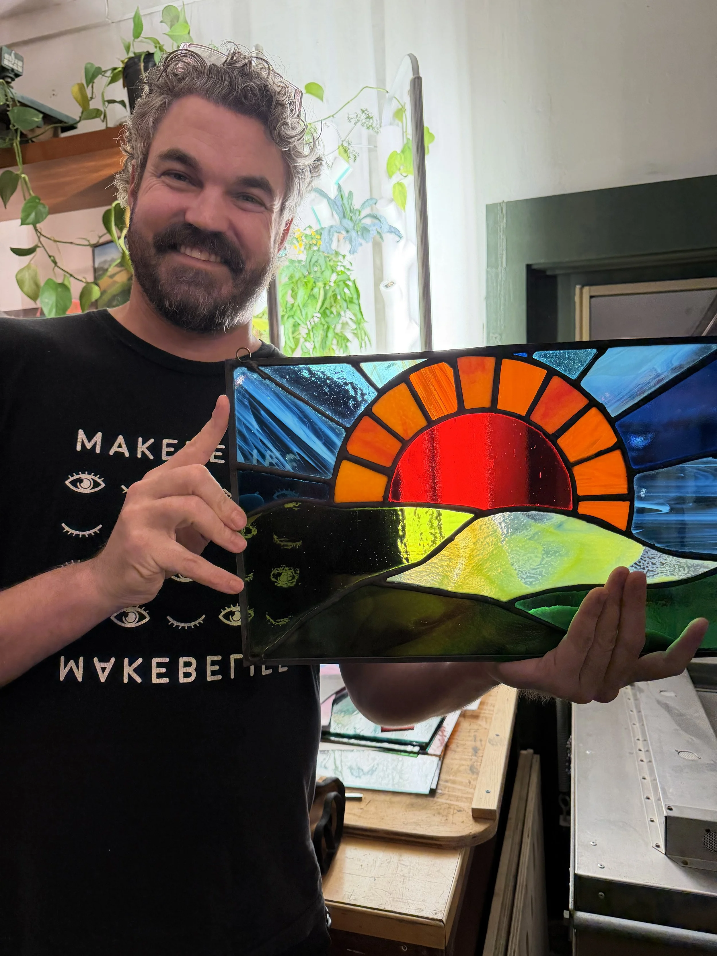 A man with gray, curly hair and a beard, smiling while holding a colorful stained glass art piece in a workshop filled with plants and workbenches.
