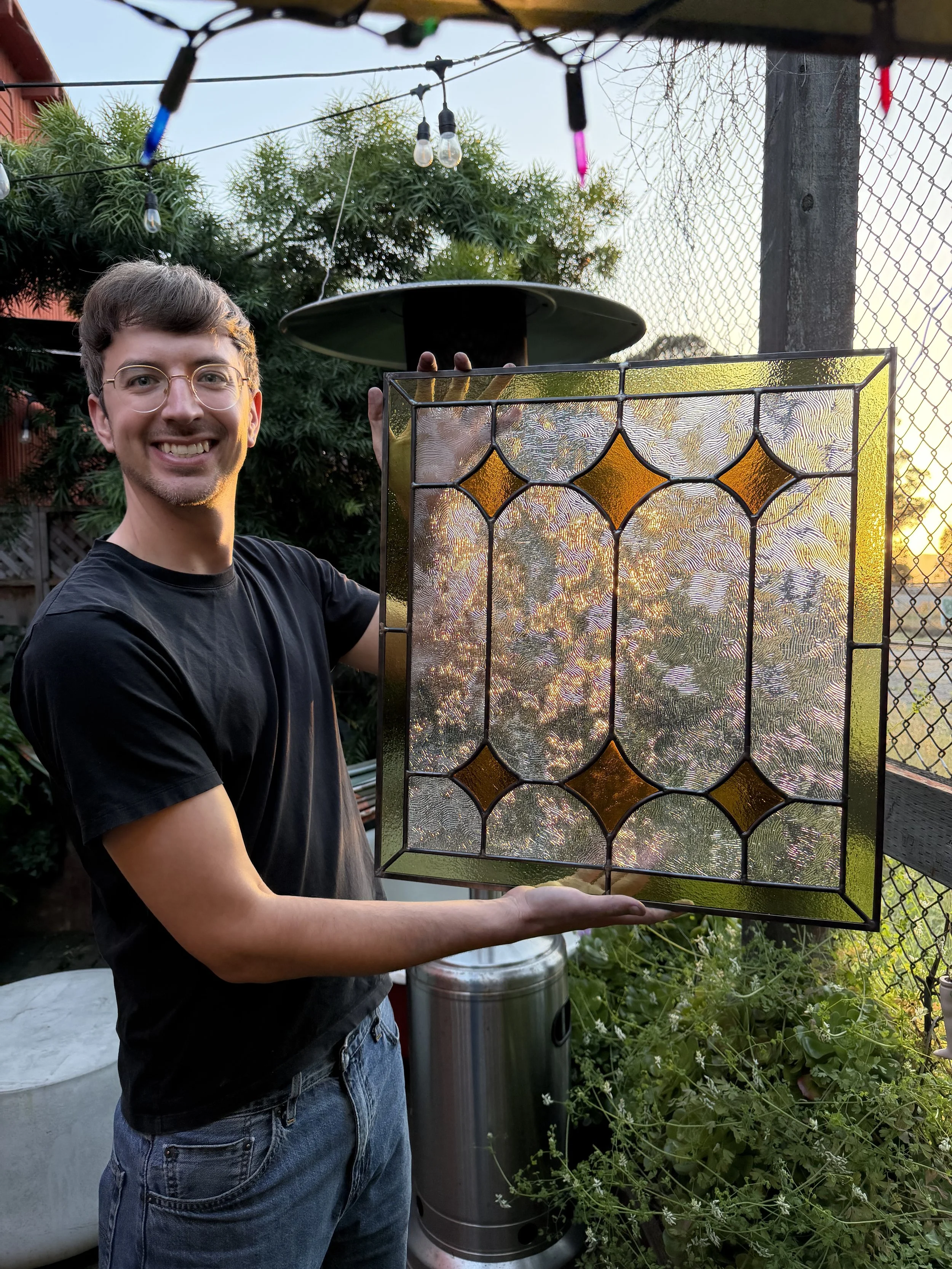 A young man with glasses smiling and holding a stained glass art piece outdoors, with string lights, trees, a fence, and a metal trash can in the background during sunset.
