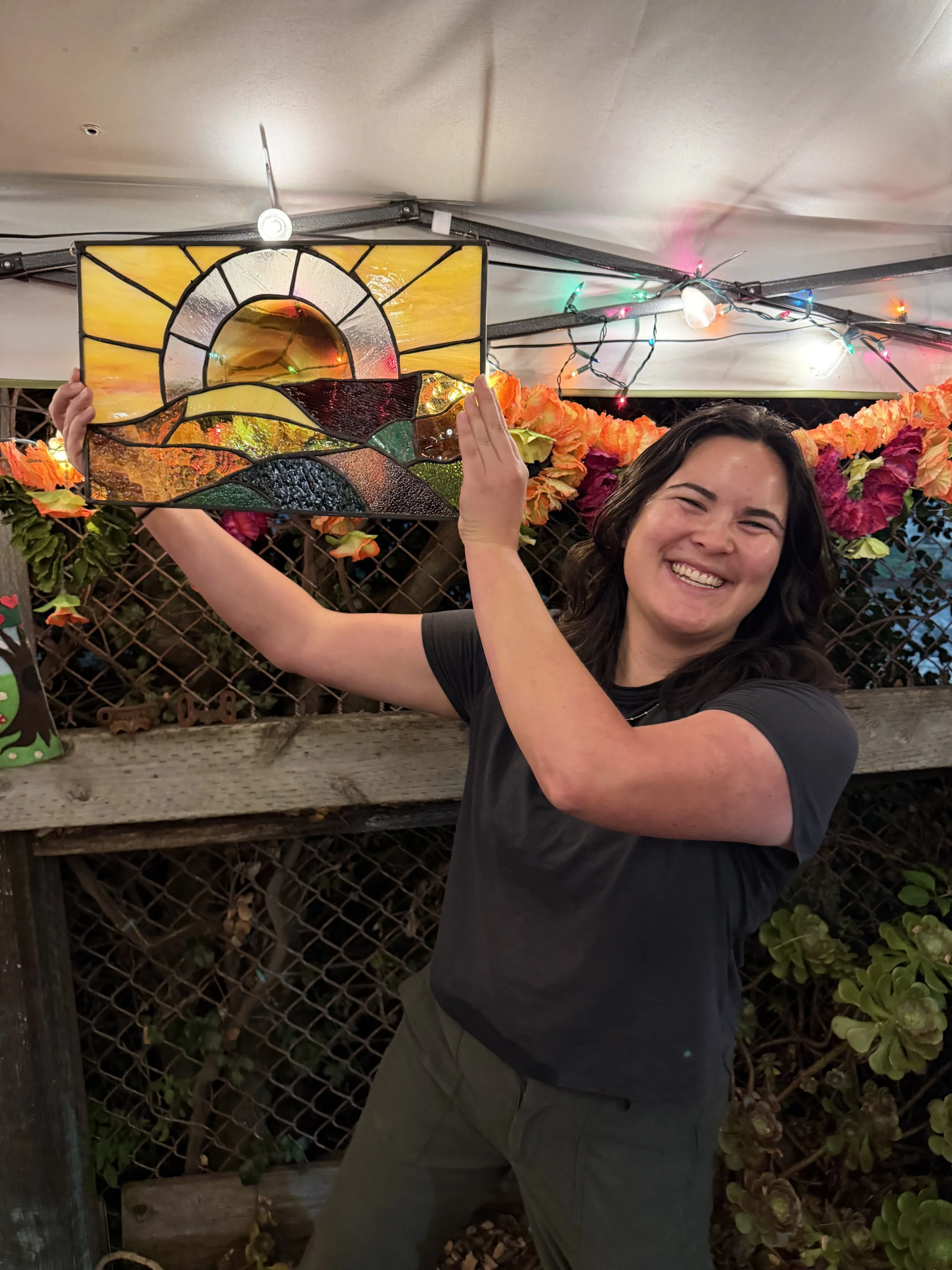 A woman with dark hair smiling and holding a colorful stained glass piece in front of a fence decorated with orange and pink flowers and string lights.