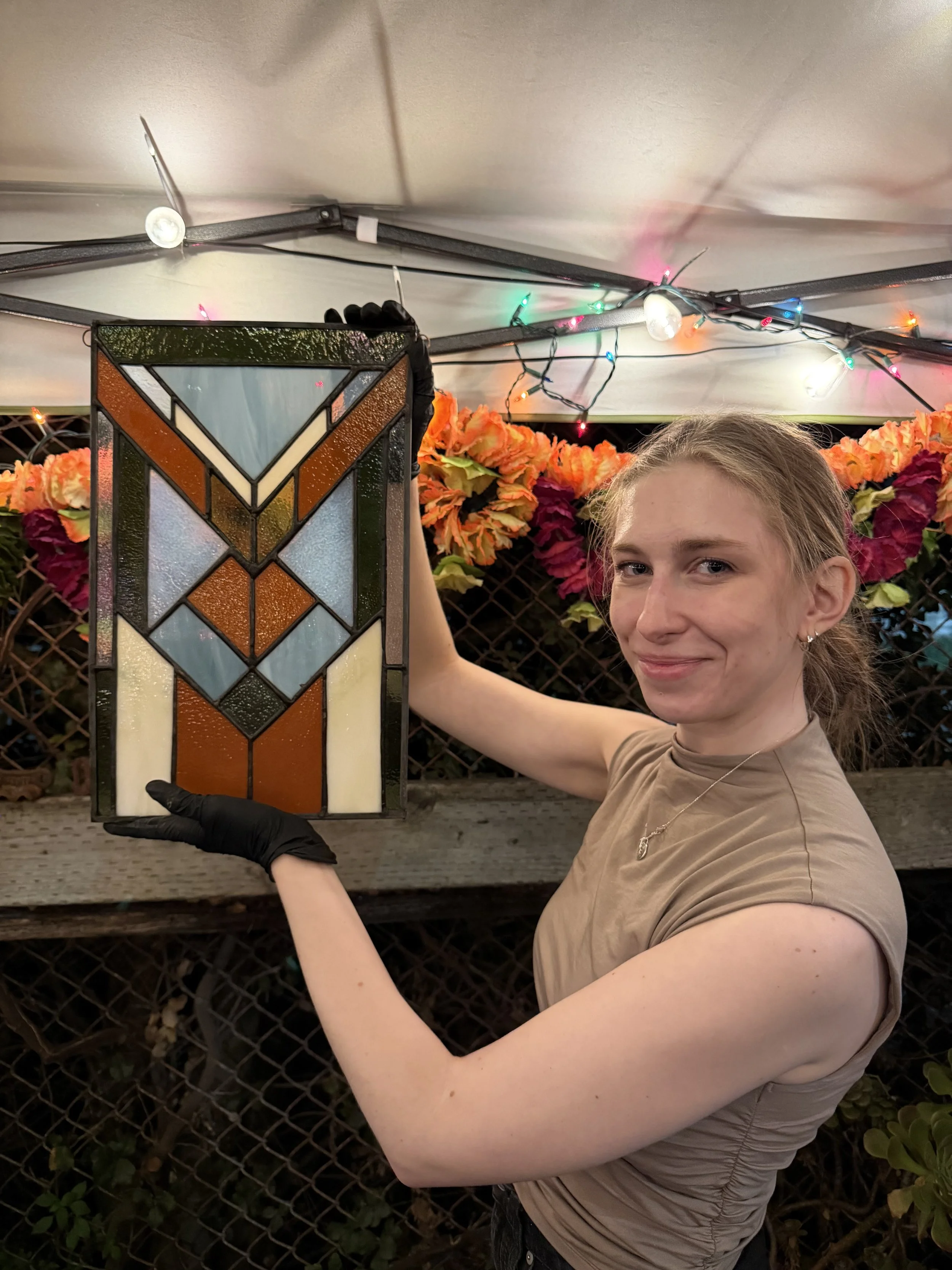 A young woman holding a stained glass lantern at night, standing outdoors in front of a flowered fence with colorful string lights overhead.