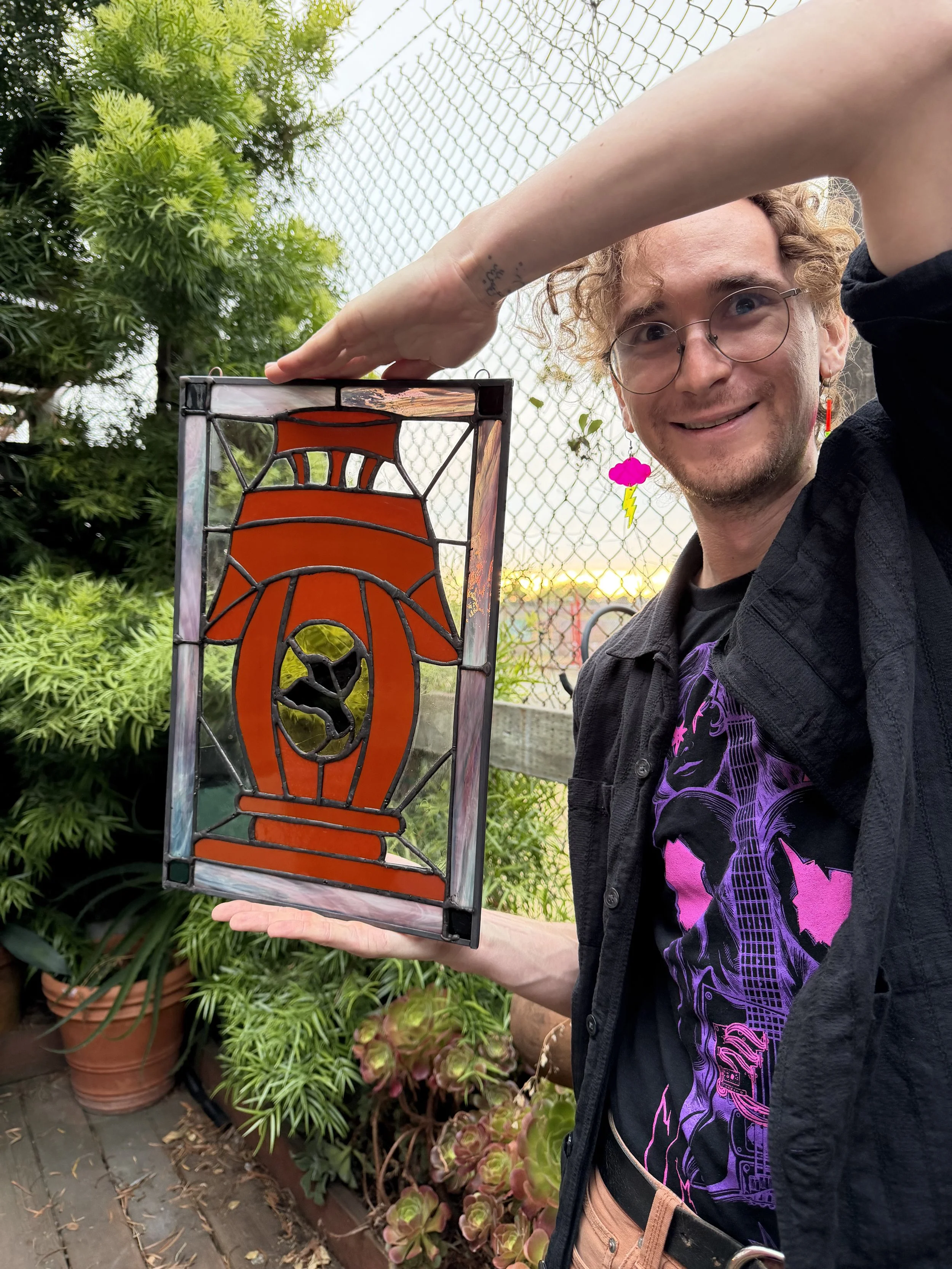A person with curly hair and glasses holding a stained glass artwork of a flaming torch with a sunset background.