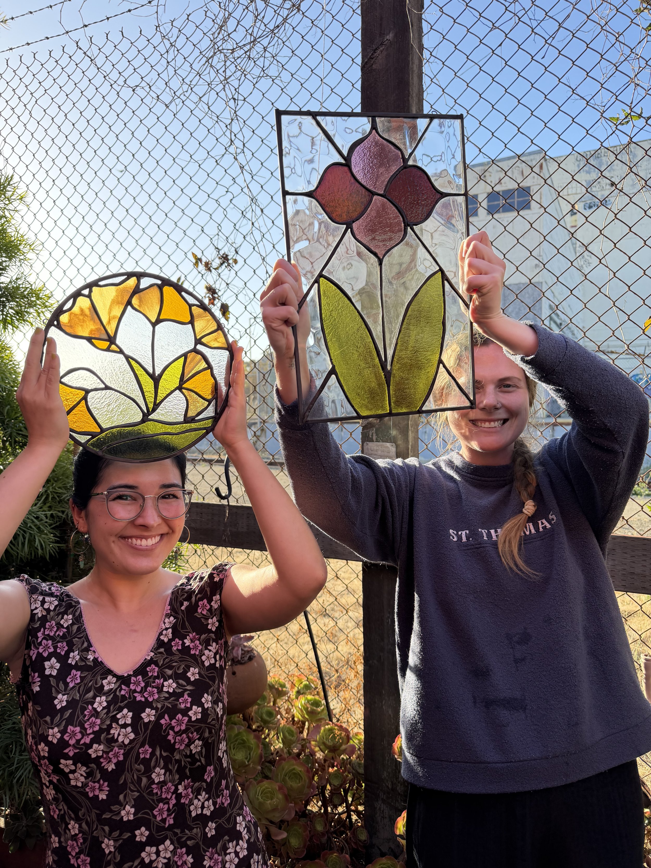 Two women holding stained glass art pieces outdoors against a chain-link fence with a blue sky in the background.