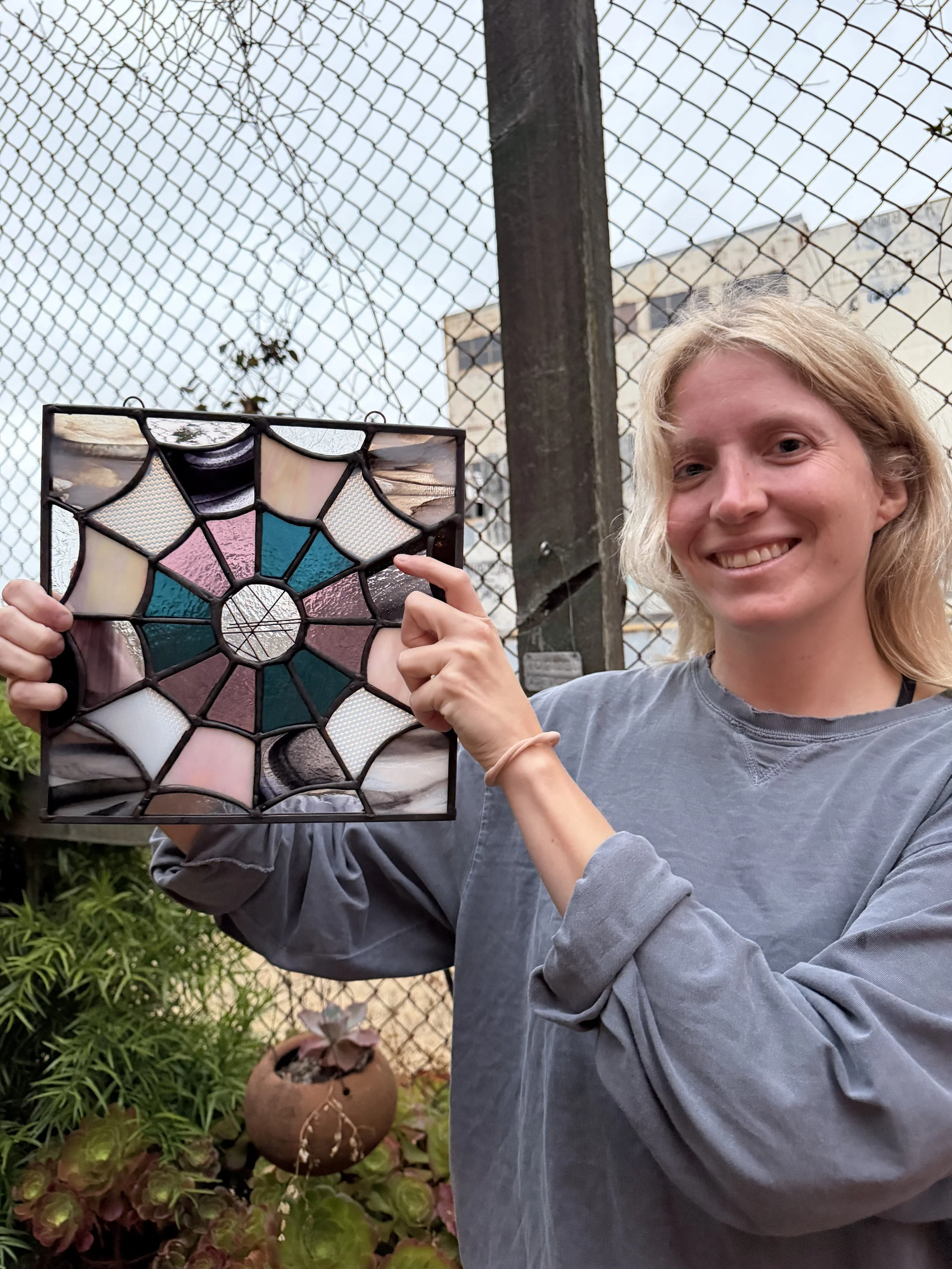A person smiling and holding a stained glass artwork in front of a chain-link fence with plants and buildings in the background.
