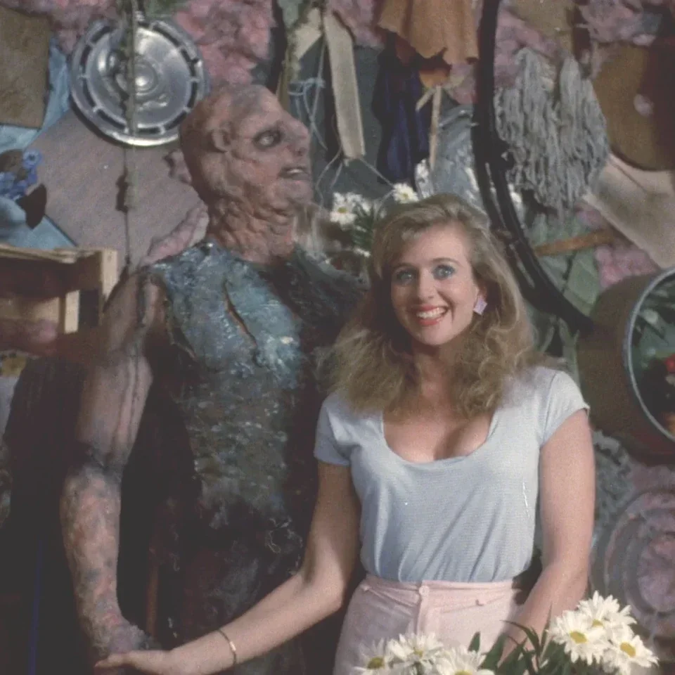 A woman with blonde hair and a woman-like monster with decayed skin and tattered clothes standing together in a cluttered room with various objects hanging on the wall.