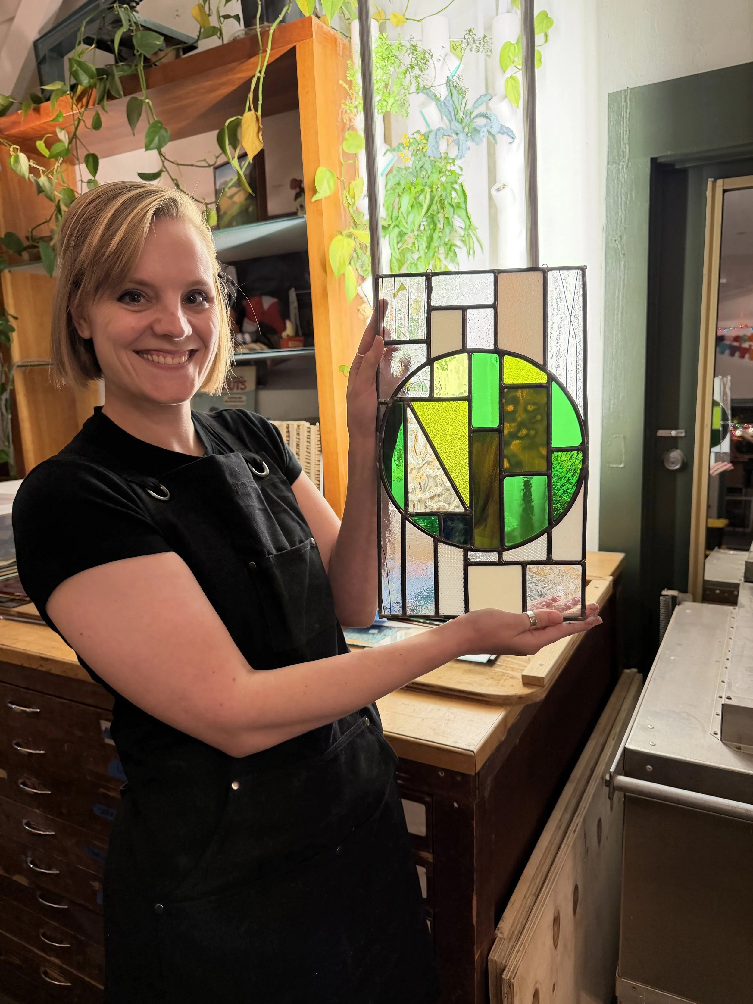 A woman smiling and holding a stained glass art piece with green and clear glass sections, standing in an indoor workshop.