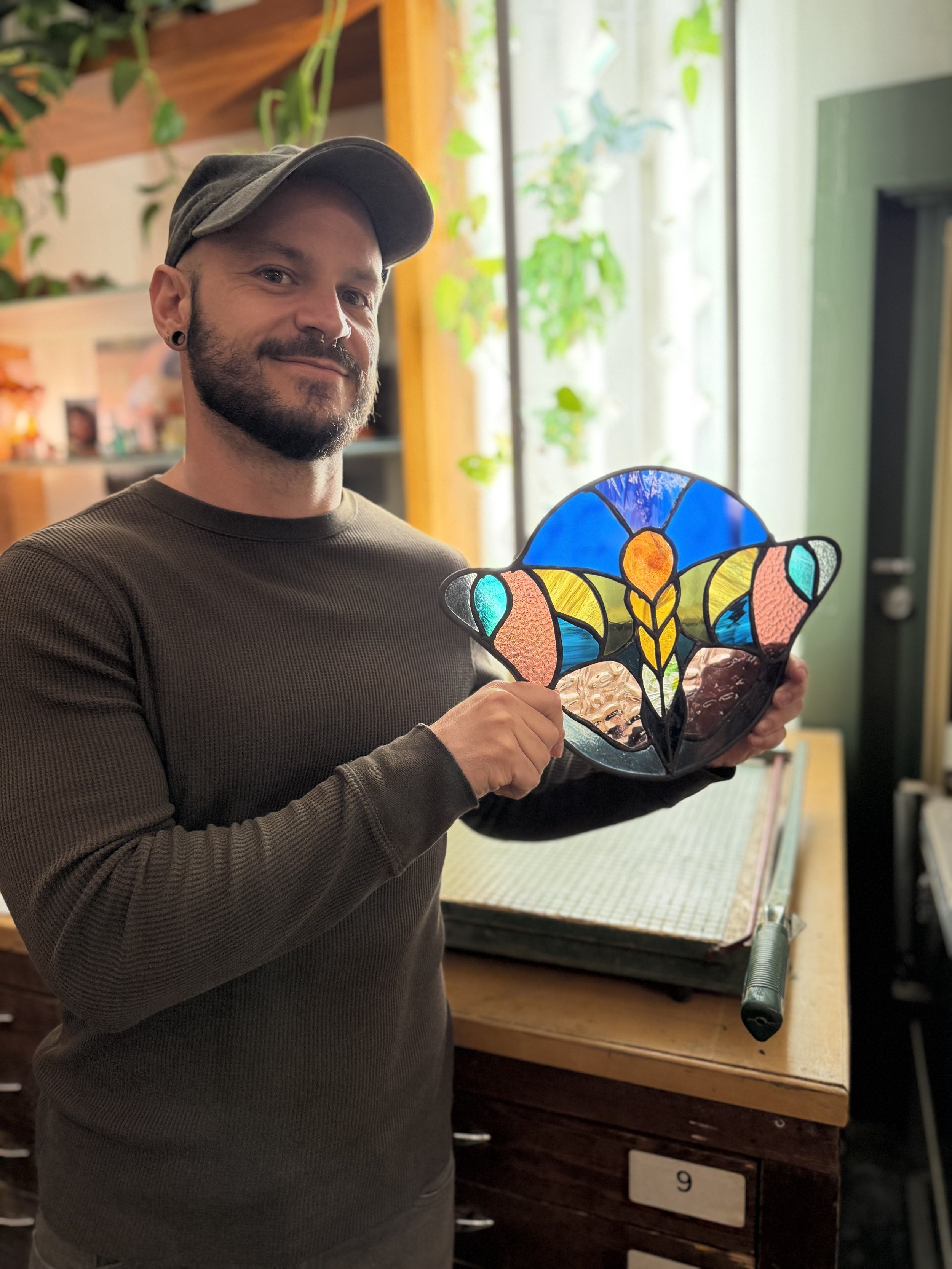 A man with a beard and dark ears, wearing a gray cap and a brown sweater, holding a colorful stained glass butterfly art piece. He is in an indoor setting with plants and wooden furniture in the background.