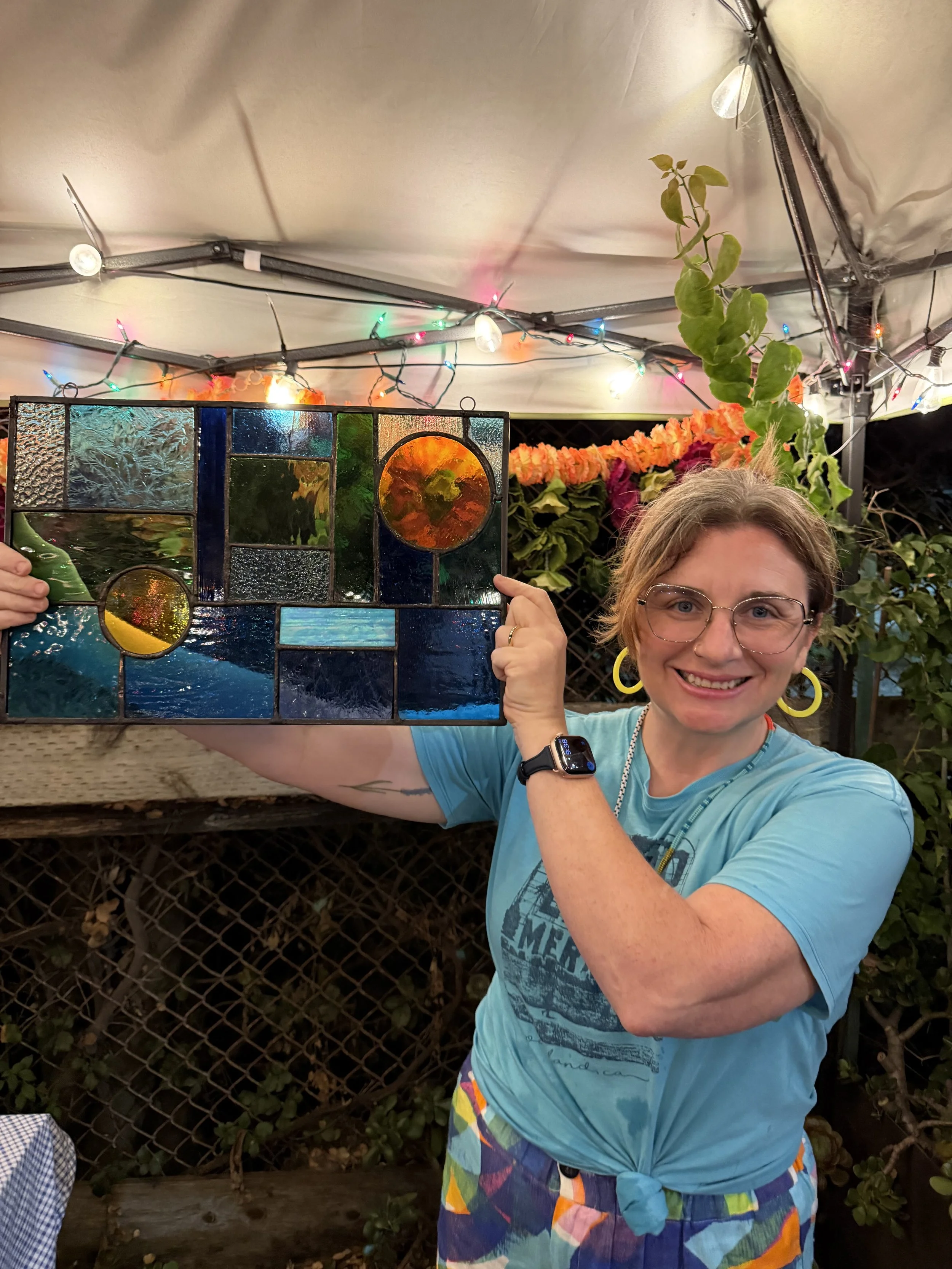 A woman with glasses and rainbow-colored shorts holds a stained glass window panel while smiling at the camera in an outdoor setting decorated with string lights, plants, and flowers.