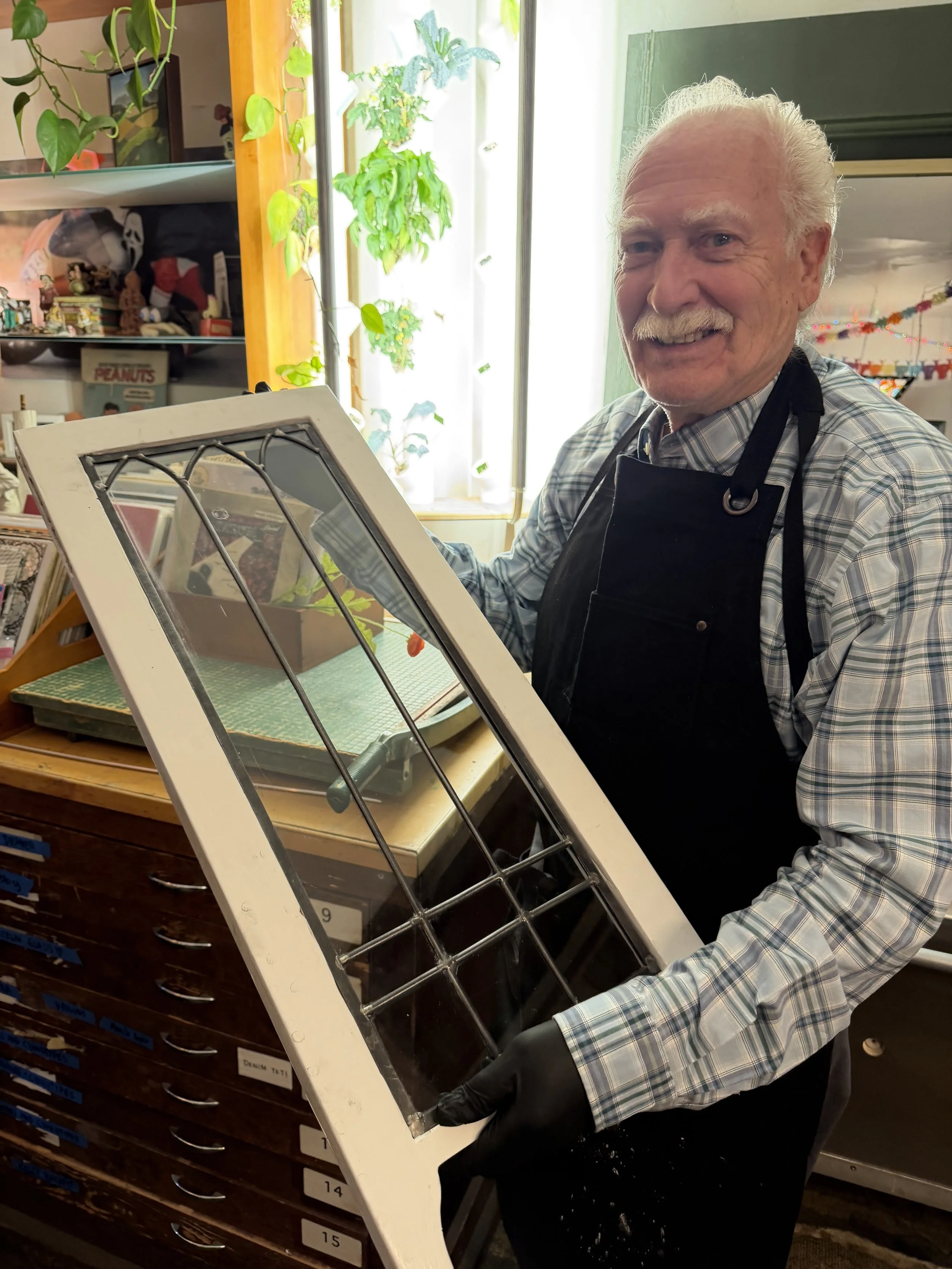 An elderly man in a plaid shirt and black apron with a smile, standing in a room with plants and Christmas decorations, holding a white canvas with a metal grid, possibly in an art or craft setting.