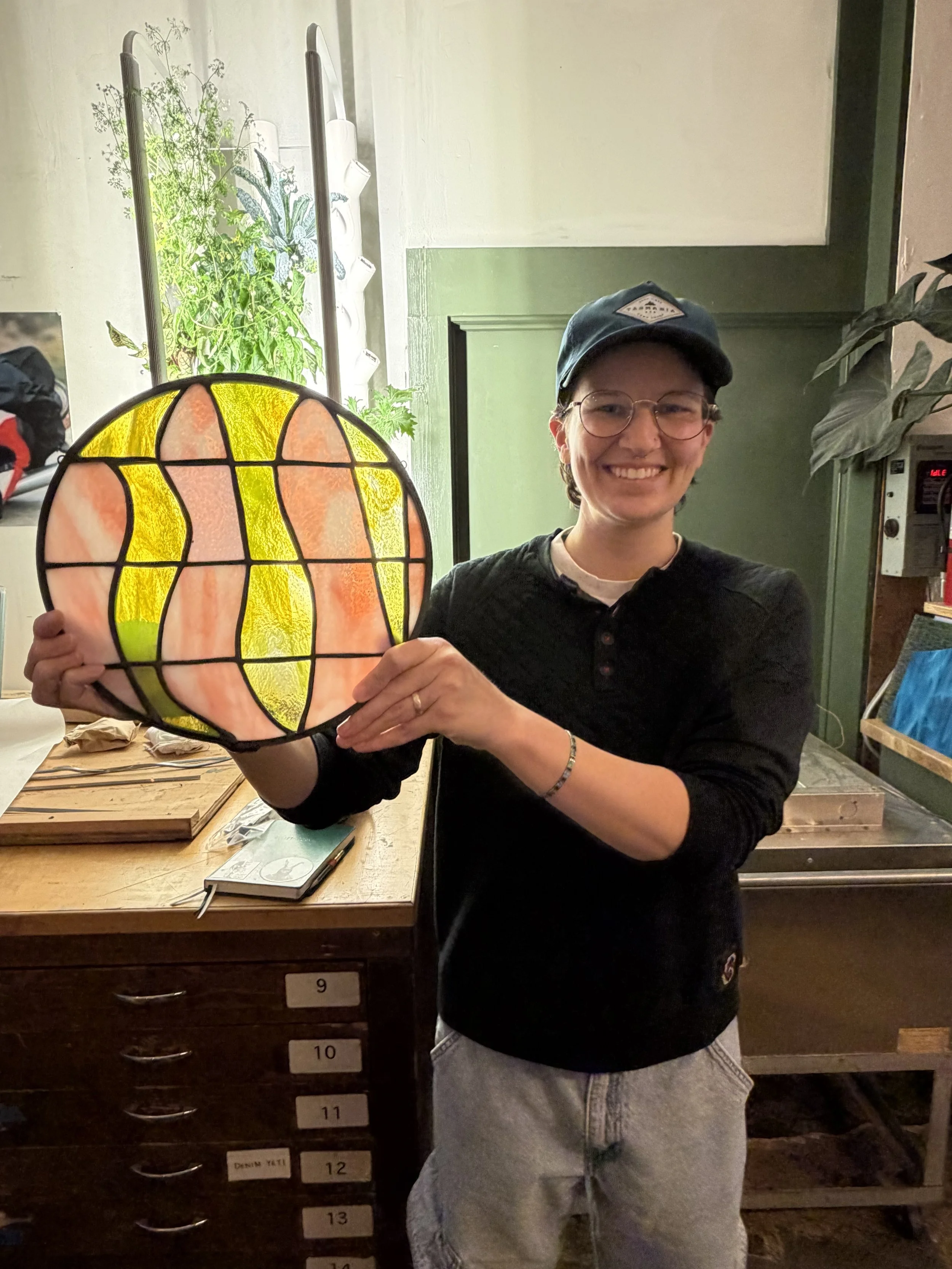A person with short hair, glasses, a baseball cap, and a black shirt, smiling and holding a stained glass art piece resembling a leaf or petal pattern with yellow and orange colors. The person is standing in front of green walls and a wooden cabinet 