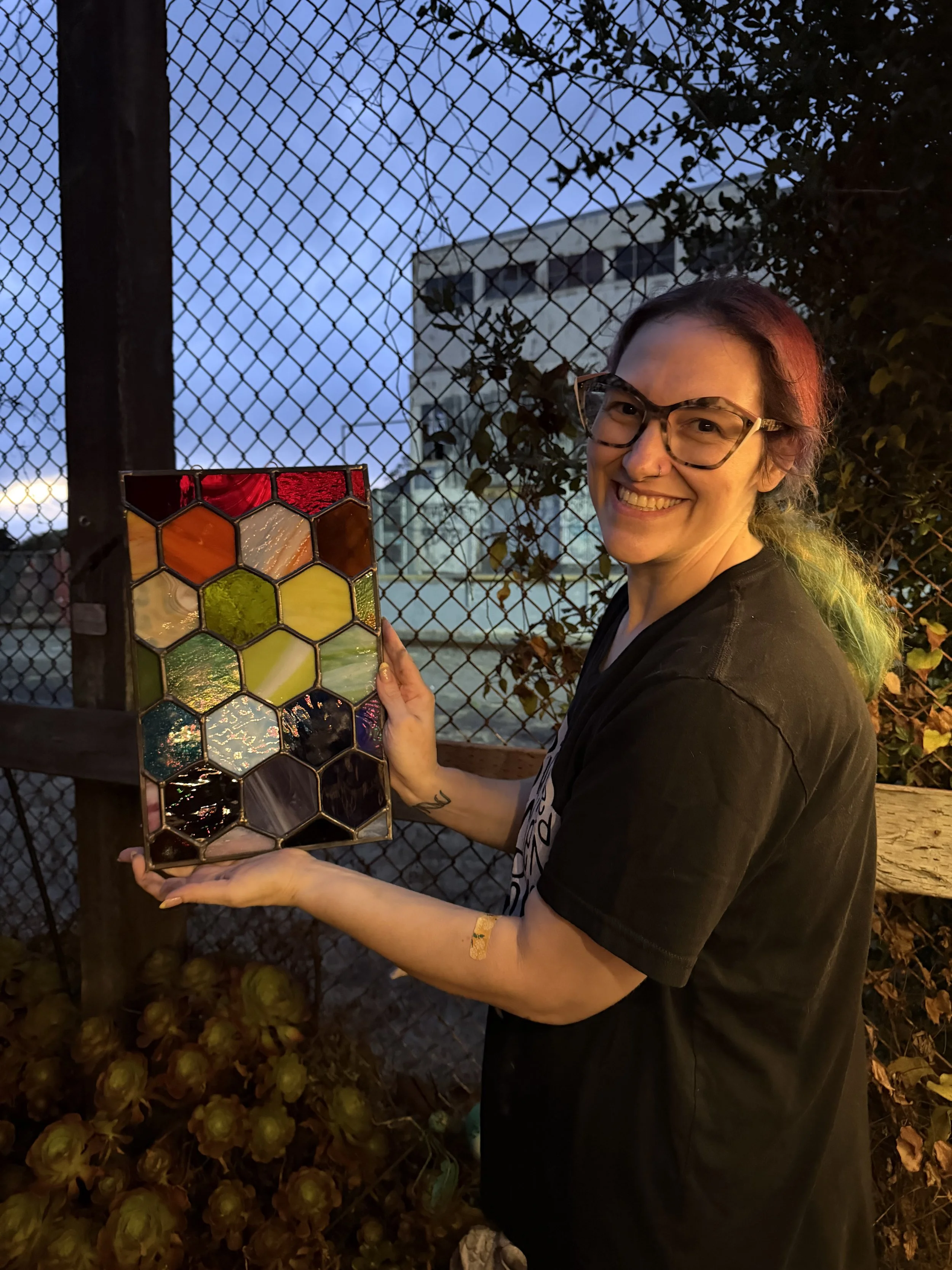 A woman with glasses smiling and holding a stained glass panel with a hexagonal pattern. She is outdoors near a wooden fence with chain-link wiring, with trees and a building in the background during dusk.