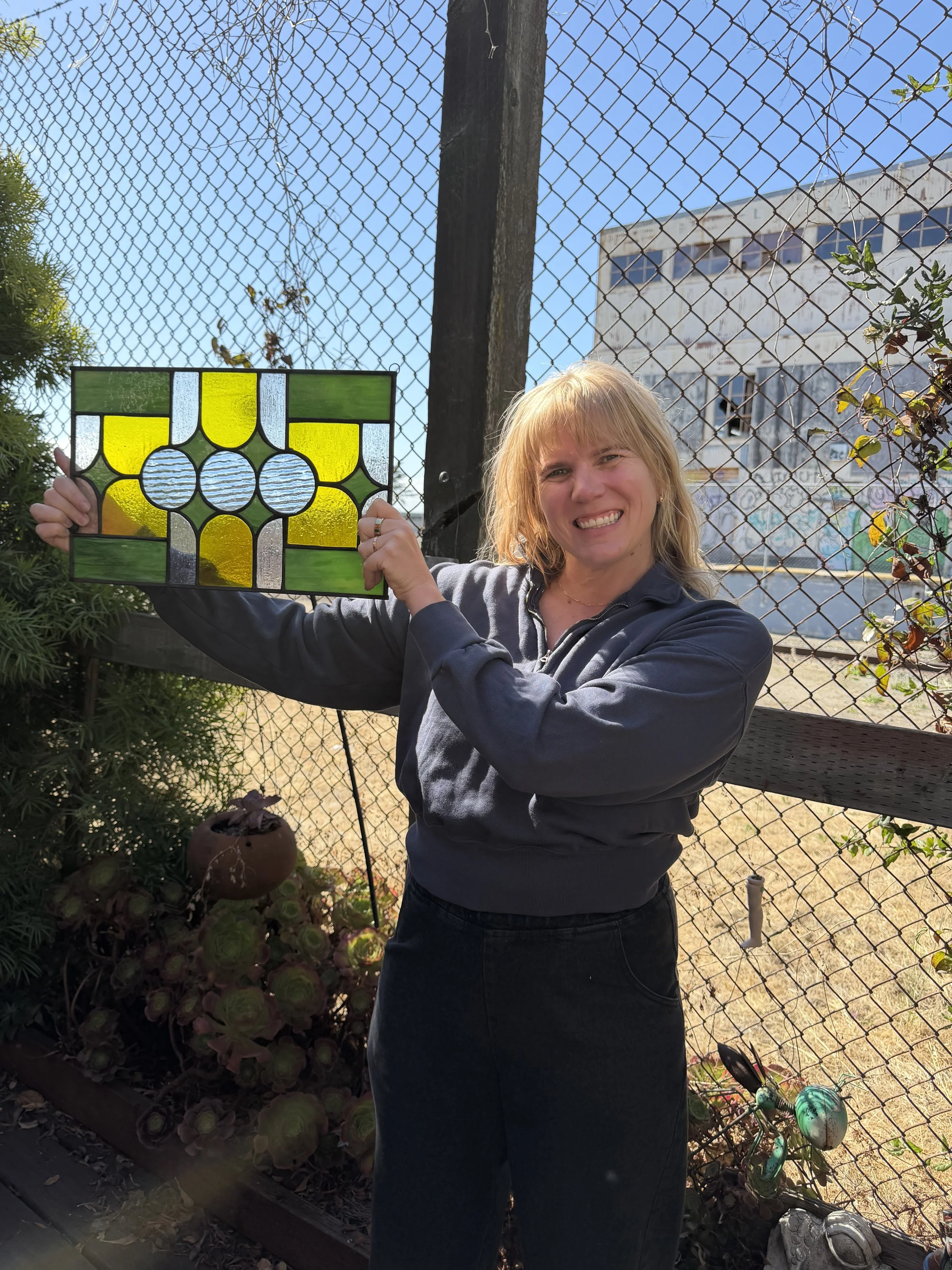 A woman smiling and holding a stained glass window panel outdoors in front of a chain-link fence, with a building and clear sky in the background.