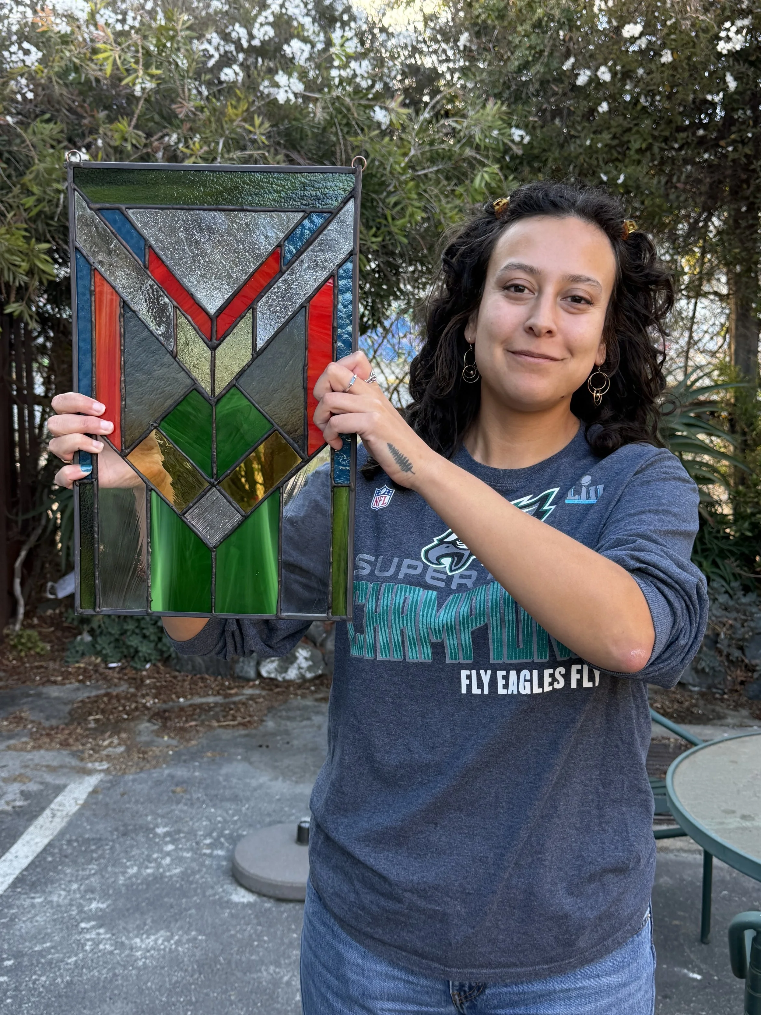Woman holding a stained glass artwork outdoors with trees and outdoor furniture in the background.