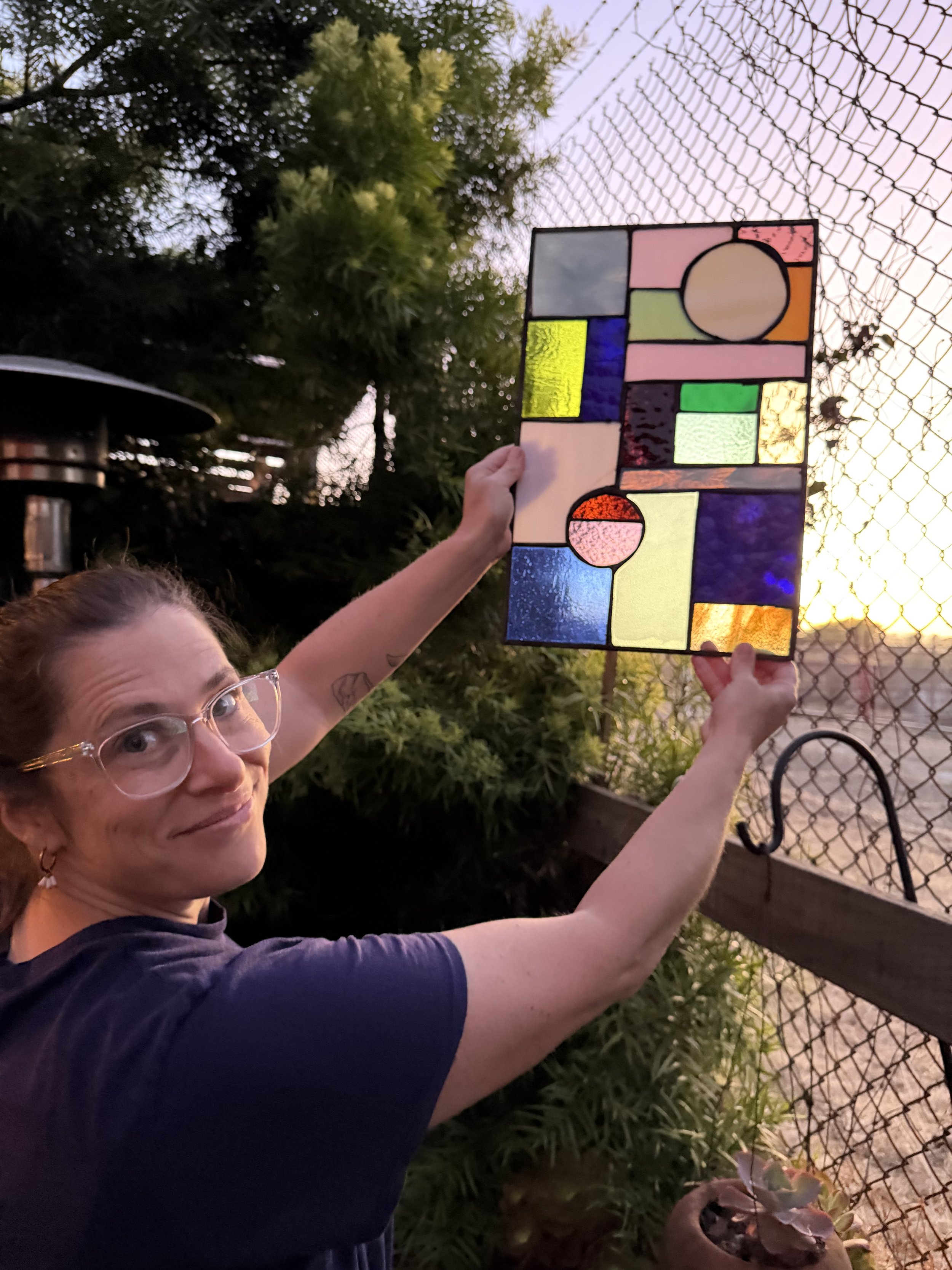 A woman holding a stained glass art piece next to a chain-link fence with trees and plants in the background at sunset.