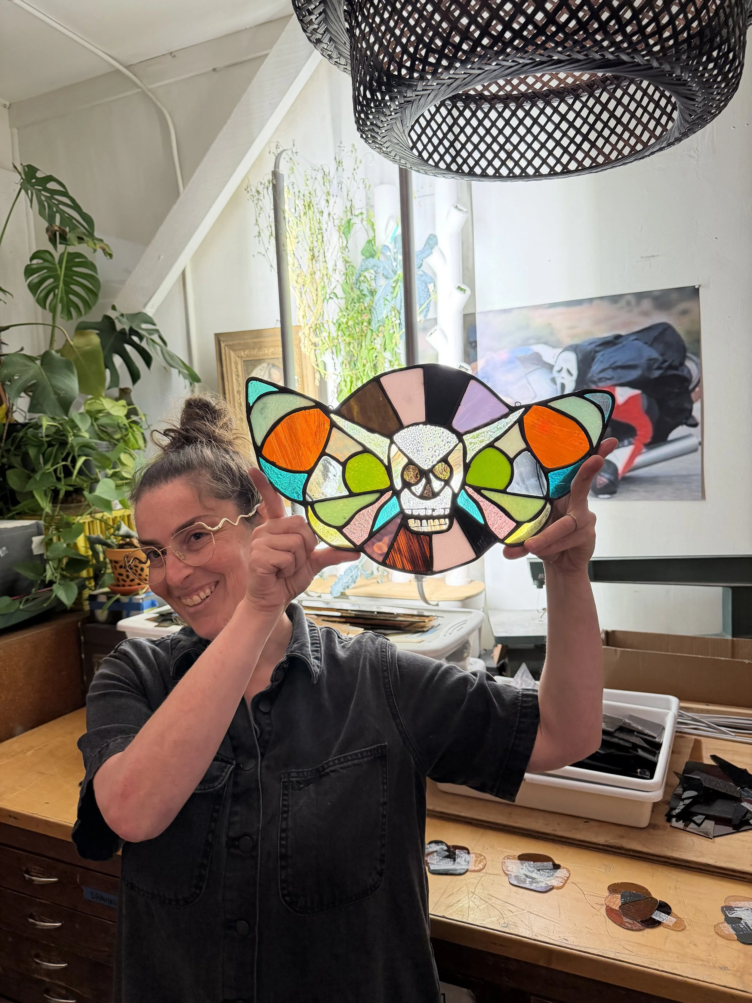 A woman smiling while holding a colorful stained glass skull lamp in front of her face. The room has plants, artwork, and a worktable with assorted stained glass pieces.