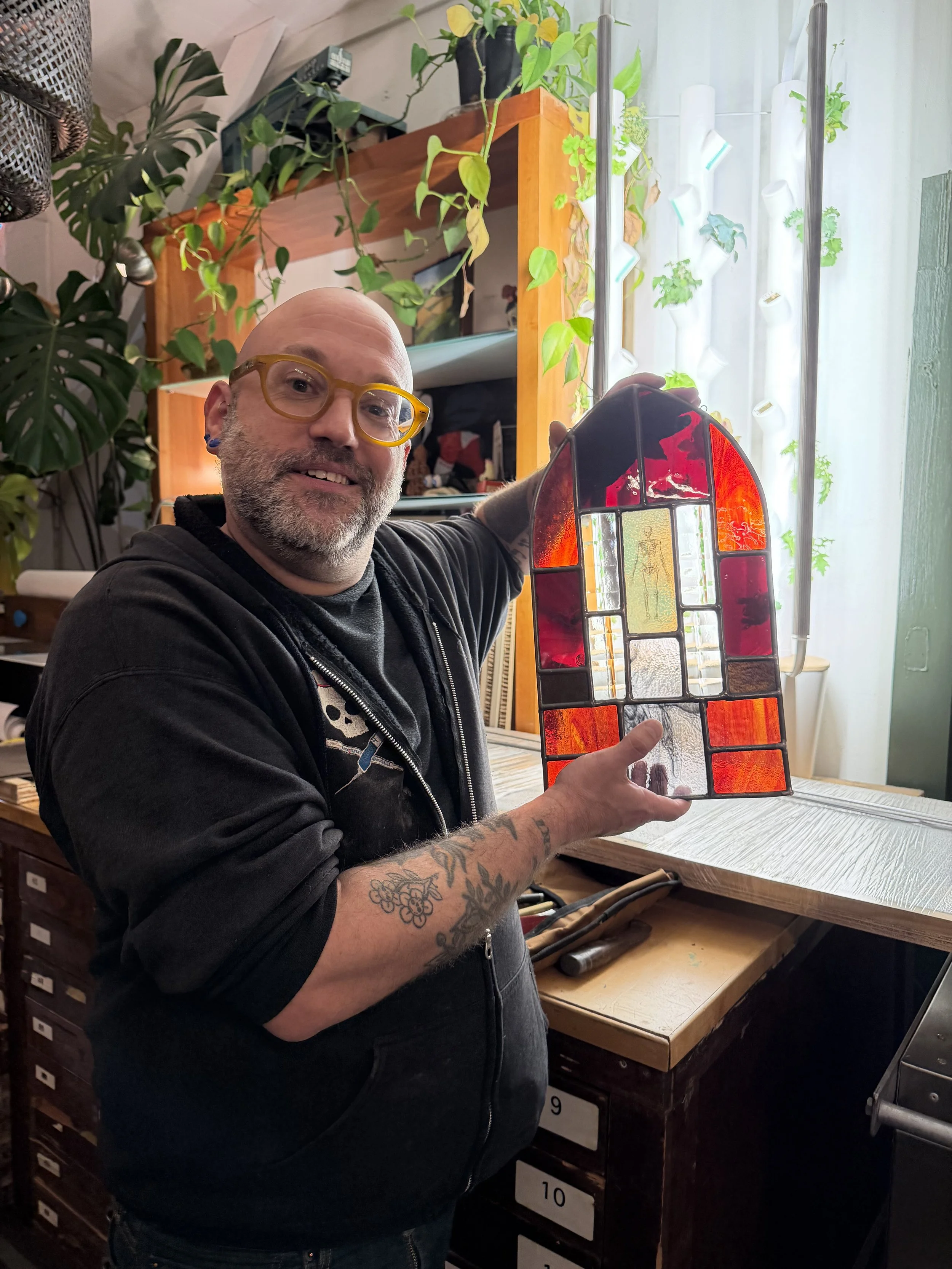 A man with glasses, a beard, and tattoos on his arm holding a stained glass art piece in a room with plants and shelves.